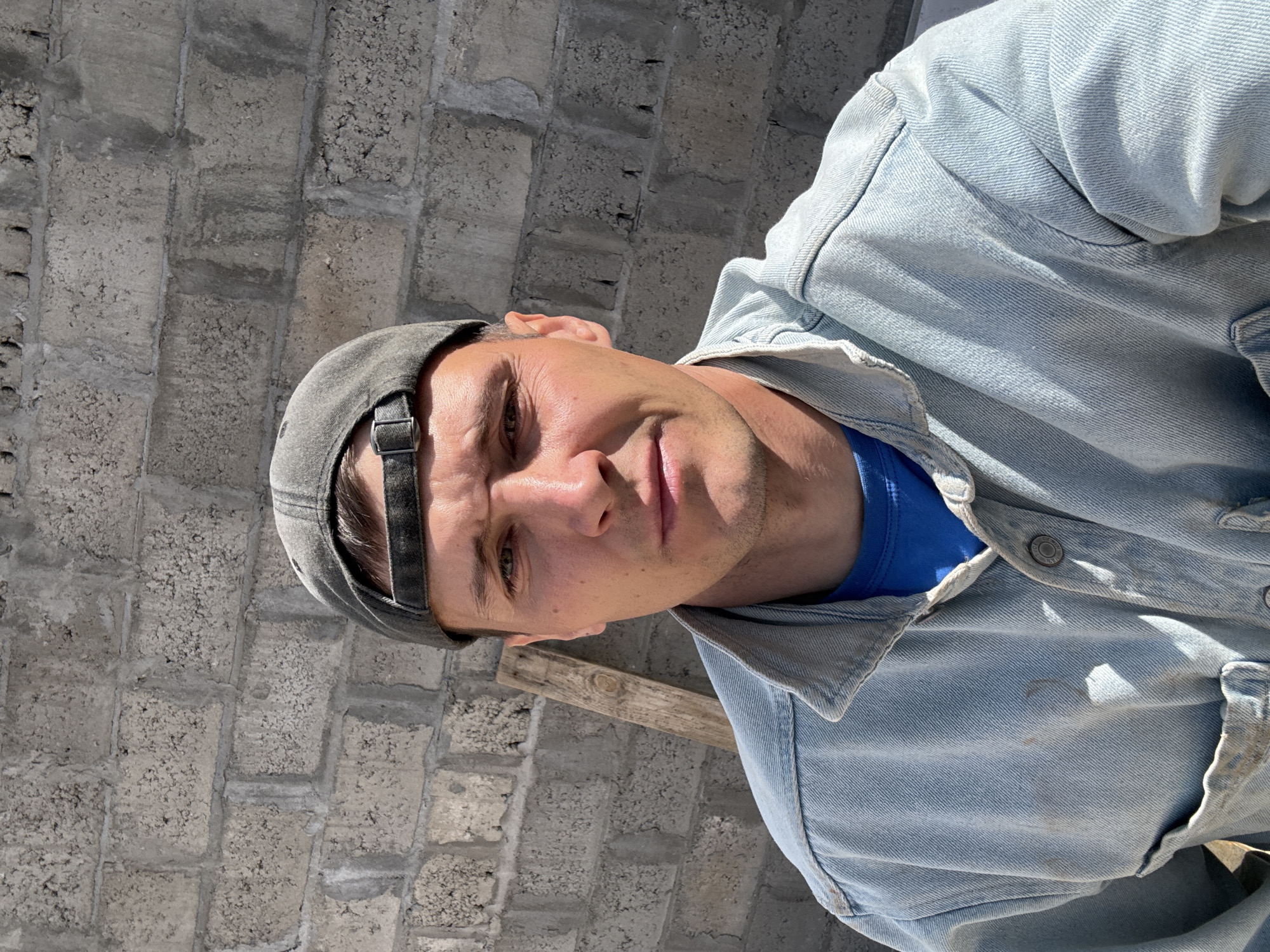 The image depicts a young adult male in a close-up, rotated 90 degrees clockwise from the original capture. He is looking upwards towards the camera, likely in a selfie. The man has dark hair, a light tan, and noticeable stubble on his face. He is wearing a dark grey or black baseball cap backward, a bright royal blue t-shirt, and a light blue denim jacket. His expression is neutral, bordering on slightly serious or thoughtful.
The background consists of a wall constructed from grey, rough-textured concrete or cinder blocks with distinct mortar lines, suggesting an unfinished or industrial setting. A wooden plank or beam is visible horizontally across the background, behind his right shoulder. The scene is brightly illuminated by direct sunlight, casting strong highlights on his face and jacket and creating clear shadows, indicating a sunny daytime with clear weather. Visible details on the jacket include seams, stitching, and metal snap buttons. No legible text is visible on the clothing or in the background. The overall setting points to an outdoor environment in Chaikovsky, Russia, possibly a construction site or an area with functional, unadorned architecture.