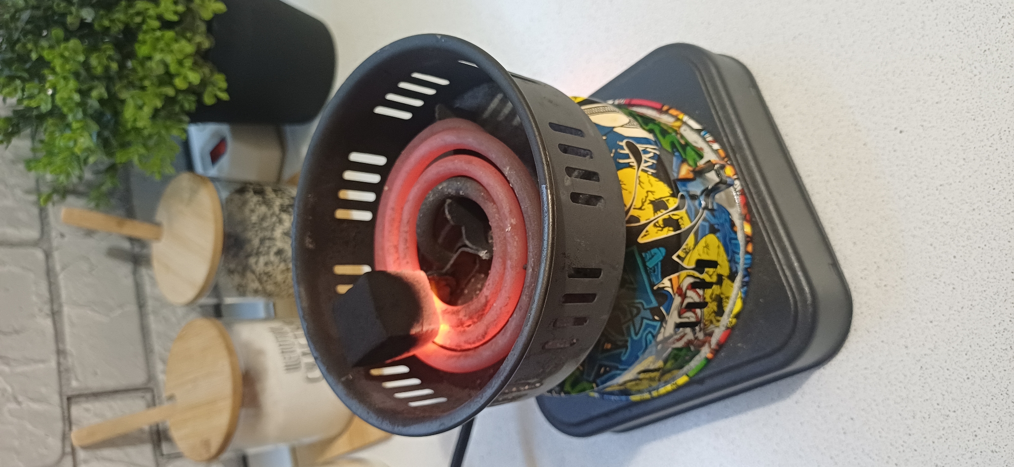 The image depicts a close-up view of an electric hookah charcoal burner. A single black charcoal cube, glowing red hot on one side, rests on the coiled heating element. The burner itself is black with slotted sides. It sits atop a colorful, graffiti-themed decorative sleeve. Below the sleeve is the black base of the burner, which has a square footprint. The background is blurred but suggests a kitchen or countertop setting with two glass jars with wooden lids, one containing a white substance and the other a granular material. A green artificial plant in a black pot is also visible to the right. The scene is well-lit. There is no indication of Zuevka, Russia, in the image.