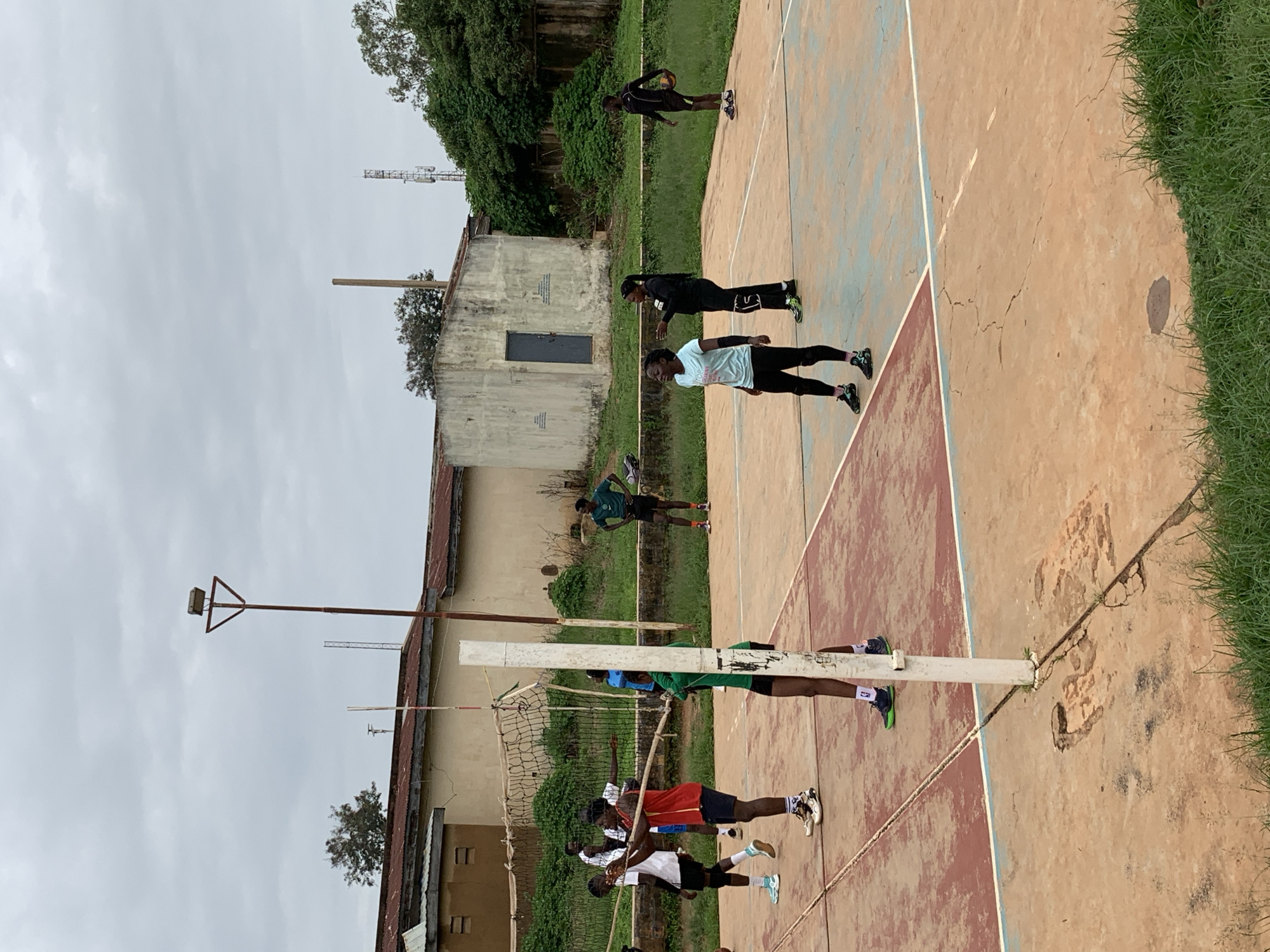 In Gura Top, Nigeria, a group of individuals are engaged in a volleyball practice on an outdoor court. The court is marked with colored lines and shows signs of wear and tear, with cracks and patches on its surface. A net stretches across the middle of the court, separating two teams. The surrounding environment includes grassy areas, buildings with corrugated roofs, and a tall light pole. The sky is overcast, suggesting a cloudy day. The participants, dressed in athletic wear, are actively involved in the game, with some preparing to serve or receive the ball. The scene captures a moment of athletic activity within a somewhat rundown, yet functional, community sports facility.