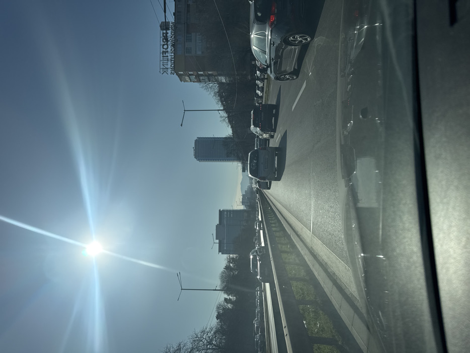 The image depicts a daytime scene on a multi-lane urban thoroughfare, identified as Tsarigradsko Shose Boulevard in Sofia, Bulgaria, looking in an easterly direction. The viewpoint is from inside a vehicle, showing the top of the dashboard and a reflection on the windshield in the foreground.
The sky is clear and light blue, with a prominent, bright sun exhibiting significant lens flare in the upper left quadrant, indicating direct sunlight and a low sun angle.
The road surface is asphalt, with multiple lanes separated by white painted lines. A metal guardrail with green vegetation beneath runs along the left side of the primary road, separating it from another parallel road or shoulder where additional vehicles are visible.
Traffic is moderate to heavy, consisting primarily of passenger vehicles (cars, SUVs, and vans) traveling away from the camera. In the right lane, a dark blue/black Hyundai compact vehicle is visible. In the middle lane, a silver/grey van is followed by a dark SUV. Numerous other vehicles are present in all visible lanes, extending into the distance.
In the mid-ground, streetlights with dual luminaires are positioned along the road. The urban environment features buildings on both sides of the boulevard. On the left, a modern office building with a glass facade is visible. On the right, further in the background, stands the tall, multi-story Capital Fort skyscraper. Adjacent to the Capital Fort building, to its right, is a residential or commercial building with a sign that reads "ПРОФИЛИК" (PROFILIK), appearing reversed and inverted due to reflection. Faint outlines of mountains are visible in the far background on the horizon. Bare deciduous trees line parts of the road, suggesting a season outside of summer.