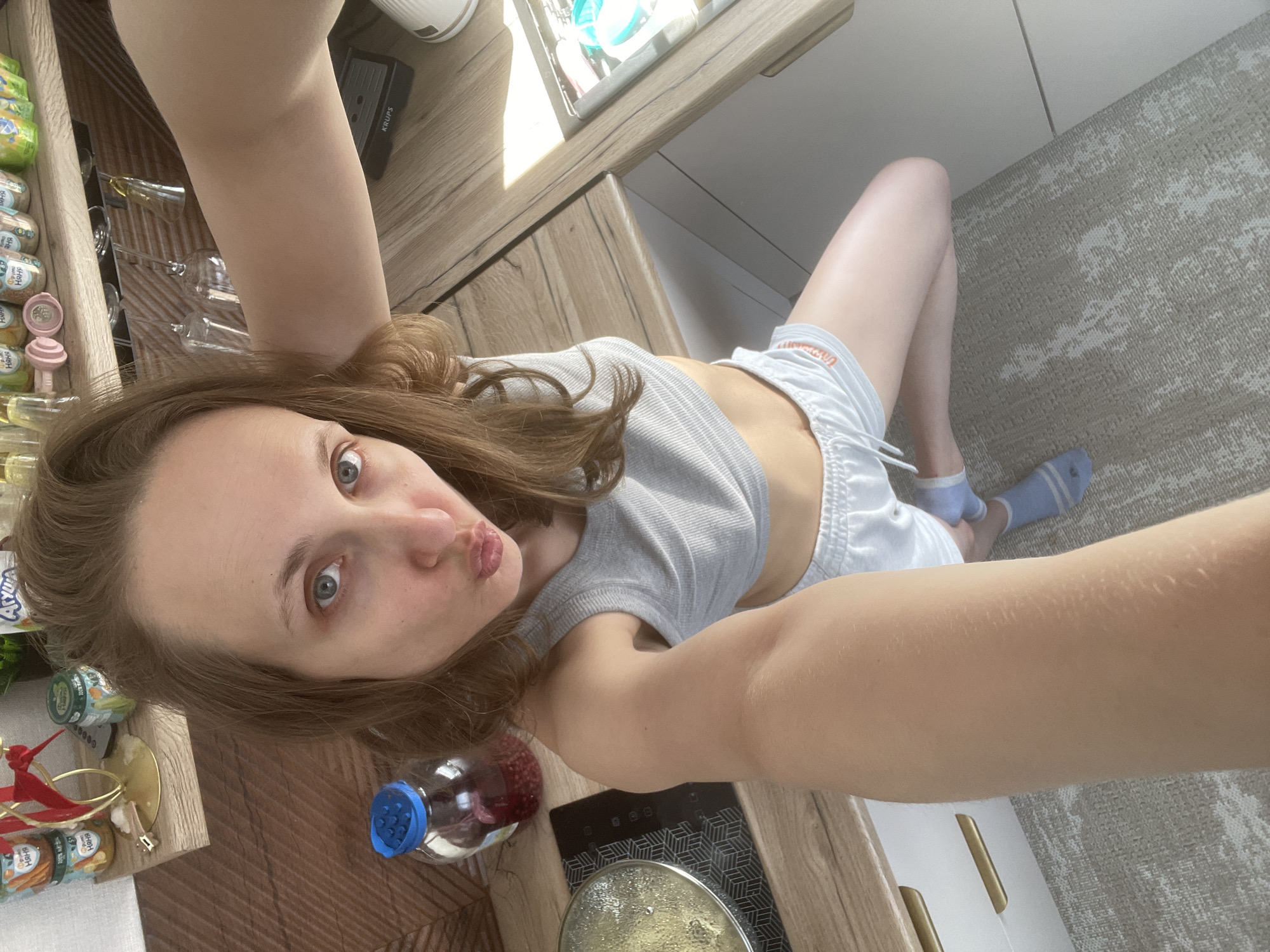 A young woman takes a selfie in a kitchen with light wood countertops, white cabinets and a gray patterned rug. She wears a gray tank top and white shorts, along with one blue sock. She is partially positioned over a black stovetop with a pot on it, and next to shelves containing a variety of goods, including baby food. The image appears to be taken from an elevated viewpoint, with the woman holding the camera or phone above her. There is a bottle with a blue cap and red contents near the stovetop.