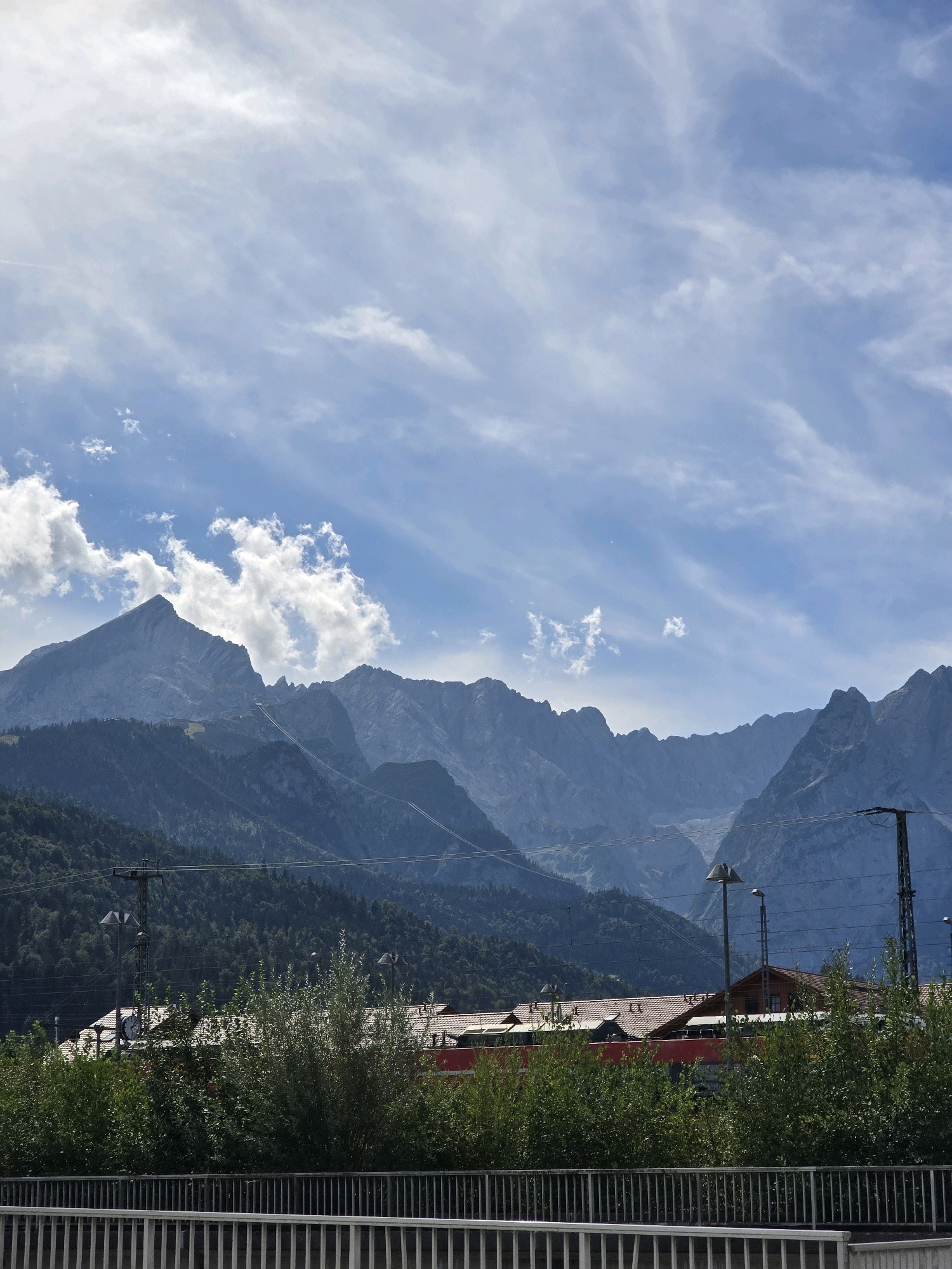 The image depicts a scenic view of the Bavarian Alps in Garmisch-Partenkirchen, Germany, under a partly cloudy sky. In the foreground, a white metal fence runs horizontally across the bottom of the frame, partially obscured by lush green foliage. Behind the foliage, a building with a red roof and red accents is visible, possibly a train station or a building associated with transportation, with power lines and lampposts in front of it.
Dominating the midground and background are the majestic, jagged peaks of the Alps, their slopes covered in dark green forests. The mountains are layered, with distinct ridges and valleys. A cable car system is visible ascending one of the mountains, indicated by two parallel cables extending upwards. The sky is a bright blue, streaked with wispy white clouds, adding depth and texture to the scene. The overall impression is one of natural grandeur and a blend of human infrastructure with the natural landscape.