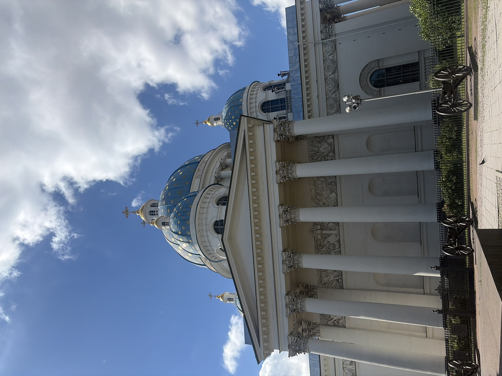 The image shows the Trinity Cathedral in Saint Petersburg, Russia, on a bright, sunny day with a partly cloudy sky. The grand cathedral features white neoclassical architecture with a prominent central dome and a smaller dome on the side, both adorned with blue and gold stars. A golden cross crowns each dome. The facade is lined with tall, fluted columns supporting an entablature decorated with bas-relief sculptures. In the foreground, two antique cannons on wheeled carriages are positioned behind a black metal fence, with a paved walkway and some greenery. The overall scene depicts a historical and architectural landmark under a clear sky, suggesting a peaceful day with minimal activity.