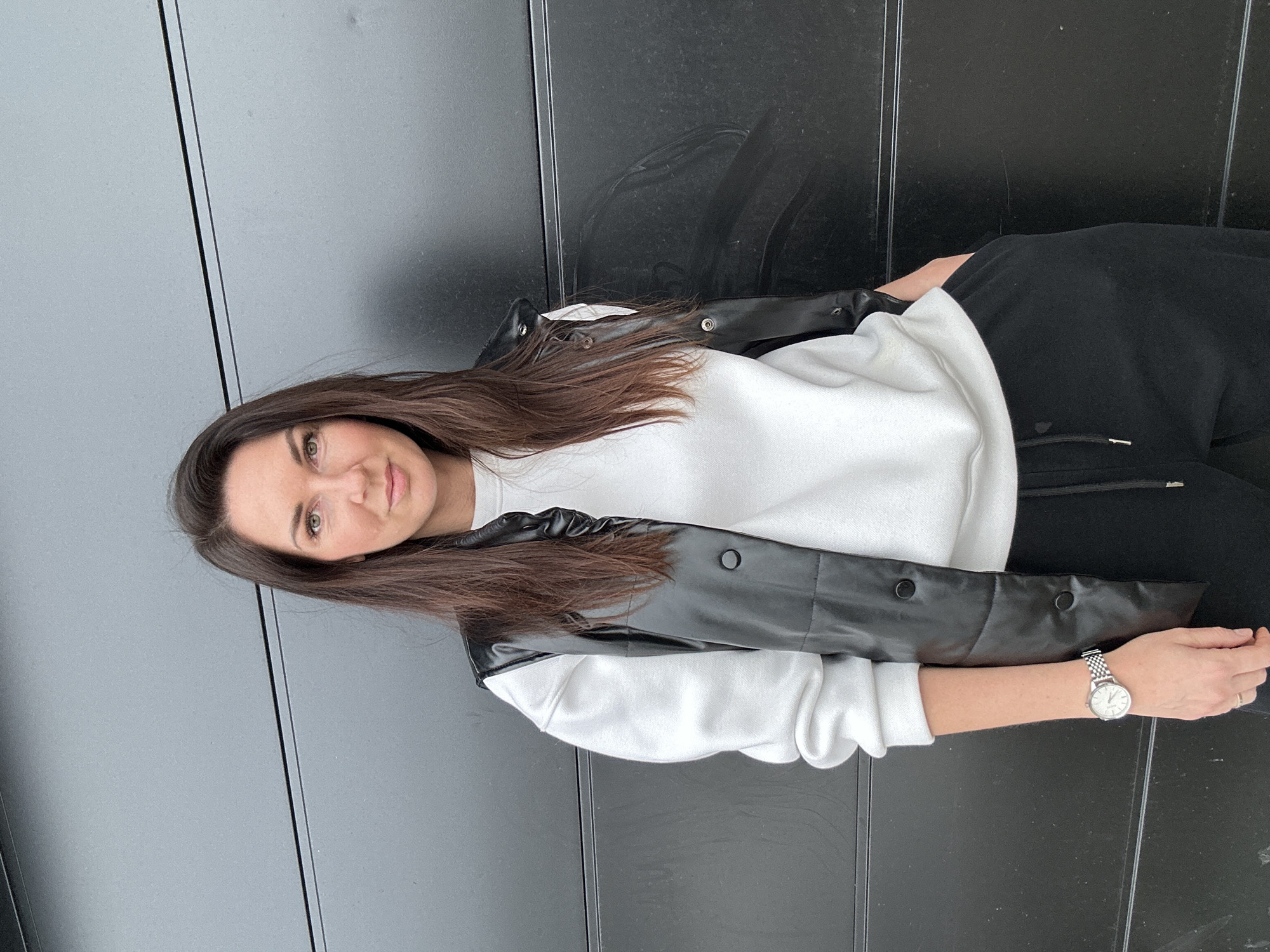 An adult female is depicted standing in front of a dark, horizontally paneled wall in Chaikovsky, Russia. The subject has long, dark brown hair parted roughly in the middle and green-hazel eyes, maintaining a neutral expression while looking directly at the camera. She is wearing a white, long-sleeved crew-neck sweatshirt, a black, shiny, sleeveless puffer vest with visible circular snap buttons, and black athletic pants or joggers with a drawstring. A silver-toned wristwatch with a round white face and a metal mesh band is present on her left wrist. Her left hand is visible at her side, while her right hand is placed into a pocket. The dark background wall, possibly an exterior garage door, exhibits horizontal seams and minor surface scuffs. The scene is illuminated by diffuse, even lighting.