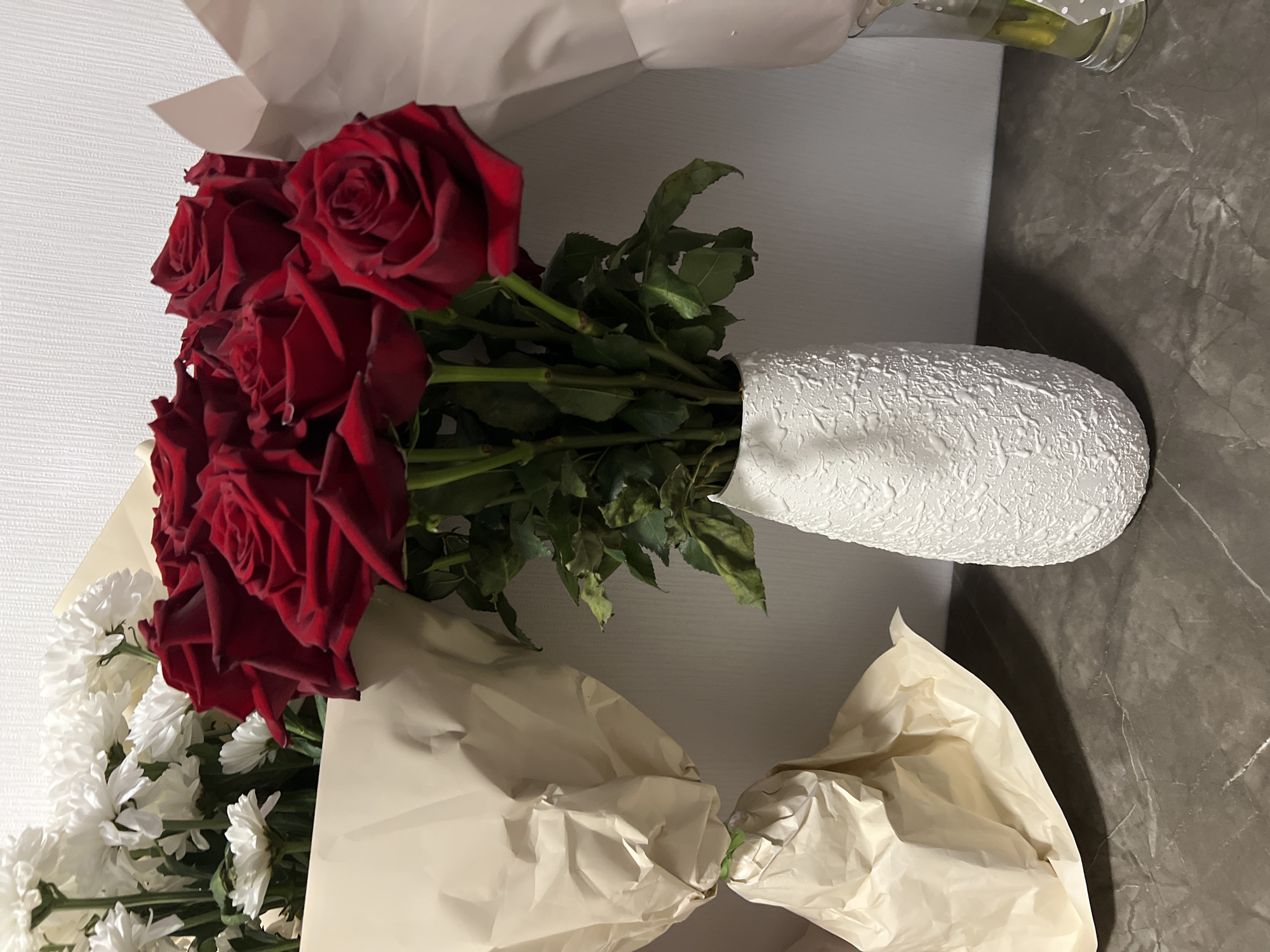 A bouquet of red roses is prominently displayed in a textured white vase on a dark, marble-patterned surface. To the left of the roses, a bouquet of white chrysanthemums is partially visible, wrapped in light brown paper. To the right of the roses, another bouquet, wrapped in a pale pink material, is also present. The background is a textured white wall. The scene appears to be an indoor setting, possibly a home or a florist shop. There is no indication of the specific location within Yekaterinburg, Russia, nor are there any people or specific actions depicted. The overall mood is one of floral arrangement and display.