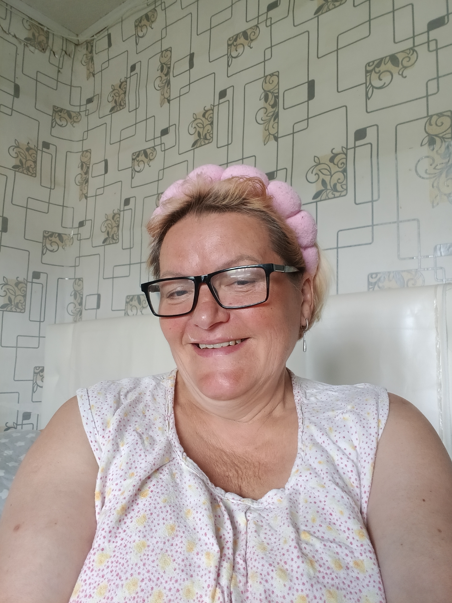 A woman with short, blonde hair and black-framed glasses smiles at the camera. She is wearing a pink, padded headband and a white nightgown with pink and yellow floral patterns. An earring dangles from her left ear. Behind her is a headboard covered in clear plastic. The wallpaper is patterned with geometric shapes and floral designs. While the location of Красный Маяк, Russia, cannot be definitively confirmed in the image, there is nothing to contradict that possibility.