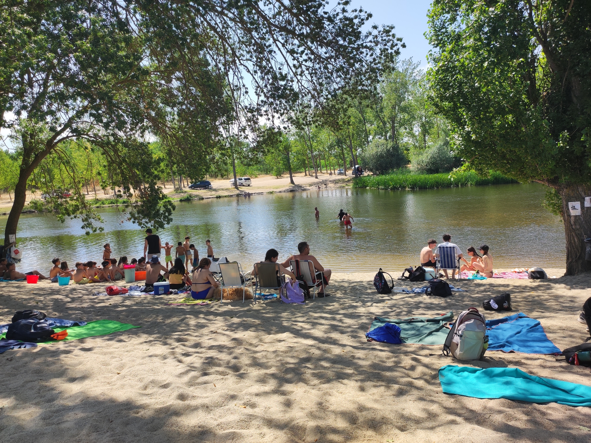 The image depicts a sunny day at a sandy beach next to a calm river. Several groups of people are relaxing on the sand and wading in the shallow water. In the foreground, beach towels and bags are scattered across the sand, some casting shadows from overhead trees. Further back, groups of people are seated on chairs or on the sand, some in swimsuits. A few individuals are standing in the river, enjoying the water.
The scene appears to be a popular recreational spot, possibly a river beach. The presence of trees with dense foliage suggests a warm, possibly summer, day. The clear blue sky and bright sunlight indicate fair weather. The water is a greenish-brown color and appears relatively shallow near the shore. The location is identified as Huerta, Spain. There are no discernible emotions visible due to the distance and resolution of the people. There is some text on a sign on the tree trunk on the right side of the image, but it is illegible.