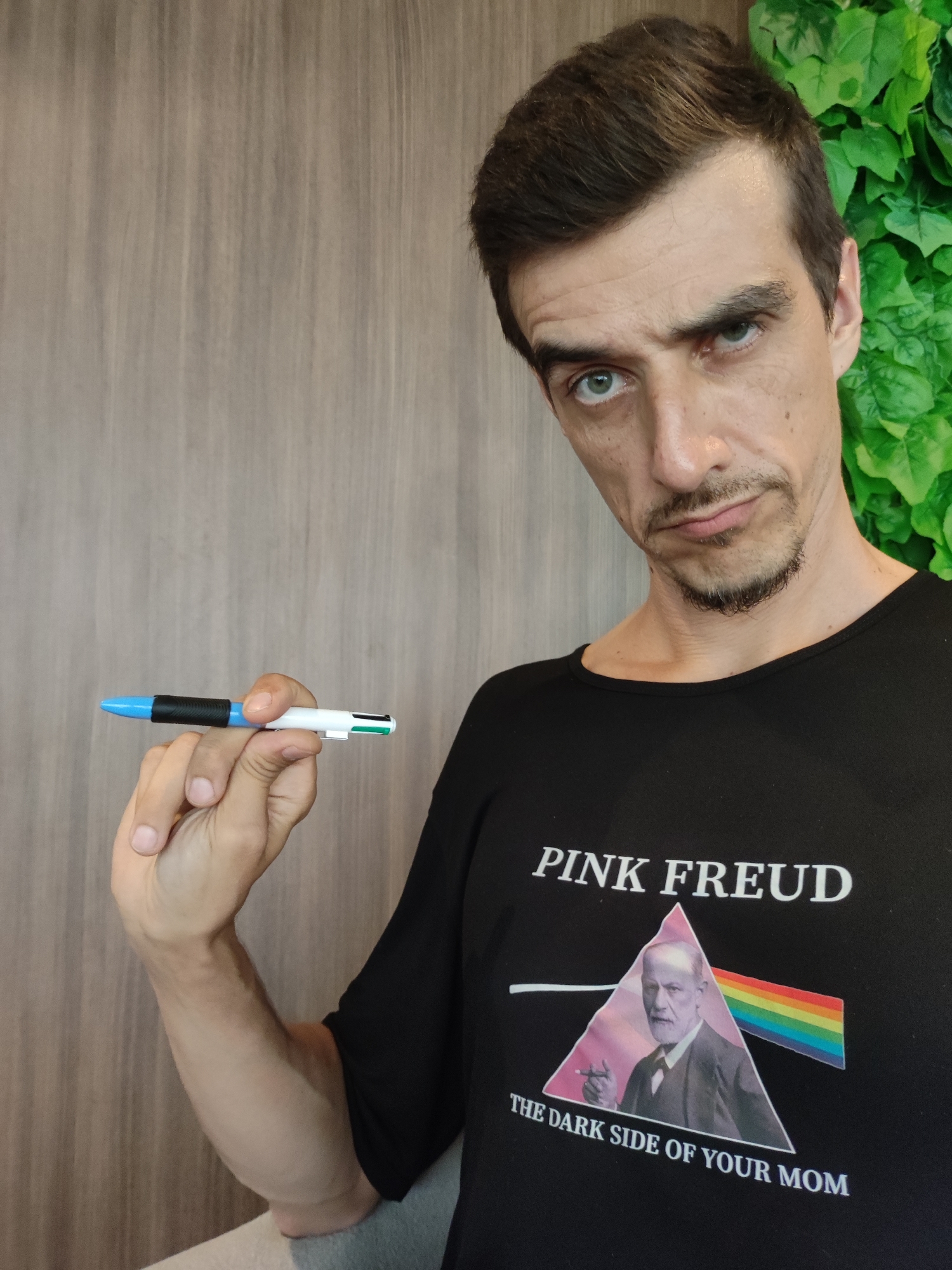 A light-skinned man with dark brown hair and a goatee looks directly at the camera with a somewhat serious or quizzical expression. He is wearing a black short-sleeved t-shirt and holds a multi-color ballpoint pen in his right hand, extended slightly forward. The pen is predominantly white and blue with black accents, featuring small green, red, black, and blue clicker buttons at its end.
The man's black t-shirt displays a graphic parodying Pink Floyd's "The Dark Side of the Moon" album cover. It features a prism-like triangle with a beam of light splitting into a rainbow. Inside the triangle is a black and white image of Sigmund Freud. Above the graphic, the text reads "PINK FREUD", and below it, "THE DARK SIDE OF YOUR MOM".
The background behind the man consists of a light brown, vertically paneled wooden wall on the left, and a wall covered in artificial green leafy plants on the right. The setting appears to be indoors or in a shaded outdoor area, likely during the daytime.
This media file was captured in North Kuta, Indonesia.