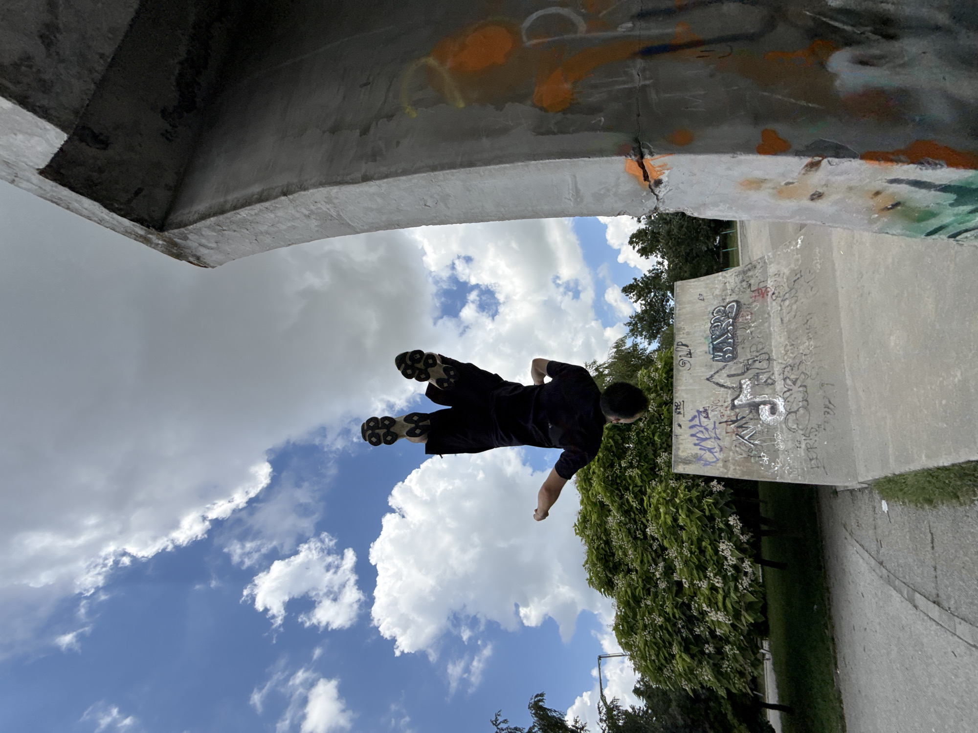The image shows a young man performing a trick on rollerblades in a skatepark. He is captured mid-air, with his body angled and his legs bent. He is wearing a dark t-shirt and dark pants, and his rollerblades are visible on his feet.
The setting appears to be an outdoor skatepark, likely in Shumen, Bulgaria, as indicated. The park features concrete ramps and structures, some of which are covered in graffiti. In the background, there are lush green trees with white blossoms and a paved pathway. The sky is a vibrant blue, dotted with fluffy white clouds, suggesting fair weather during daytime.
The scene evokes a sense of athleticism and freedom, with the subject's airborne pose highlighting the dynamic nature of the activity. The graffiti on the concrete structures adds a touch of urban culture to the environment. The image is taken from a low angle, emphasizing the height of the jump and the vastness of the sky. There is no visible text that is clearly legible.
