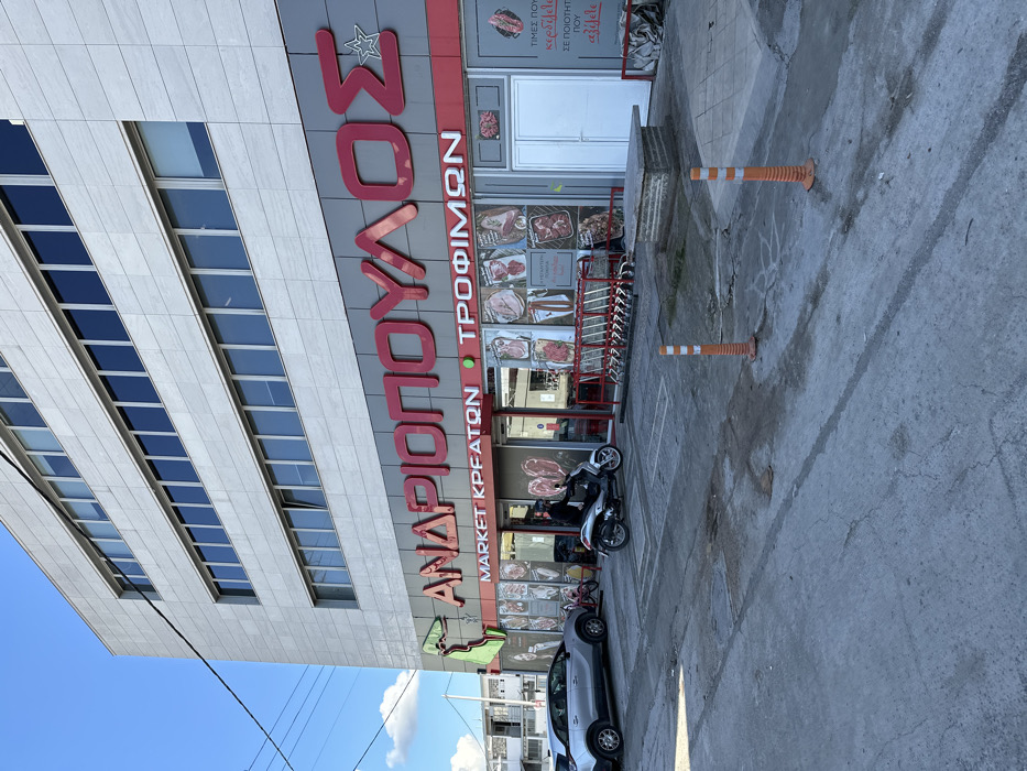 The image depicts the storefront of a commercial building, likely a meat and food market, on a bright, sunny day in Metamorfosi, Greece. The building is a modern, multi-story structure with light-colored tiled facades and numerous rectangular windows.
The ground floor features a prominent red sign with white lettering. The main name displayed is "ΑΝΔΡΙΟΠΟΥΛΟΣ" (Andriopoulos), with a star logo next to the first letter. Below it, smaller text reads "MARKET ΚΡΕΑΤΩΝ • ΤΡΟΦΙΜΩΝ" (Meat Market • Food). The storefront consists of large glass windows showcasing various meat products, such as steaks, roasts, and sausages. A small sign within one of the windows has text partially visible, including "ΤΙΜΕΣ ΠΟΥ..." (Prices that...) and "ΣΕ ΠΟΙΟΤΗΤΑ..." (in quality...). A row of red metal shopping carts is lined up near the entrance.
In front of the store is a paved, grey parking area or access road. A grey three-wheeled motorbike (scooter) is parked close to the entrance. Further to the left, a silver car is also parked. Two orange and white striped flexible traffic cones are placed in the paved area. The sky is clear and blue with a few distant clouds, indicating good weather. Some overhead power lines are visible against the sky in the lower left corner. No people are explicitly visible, but the setup suggests an active commercial environment.