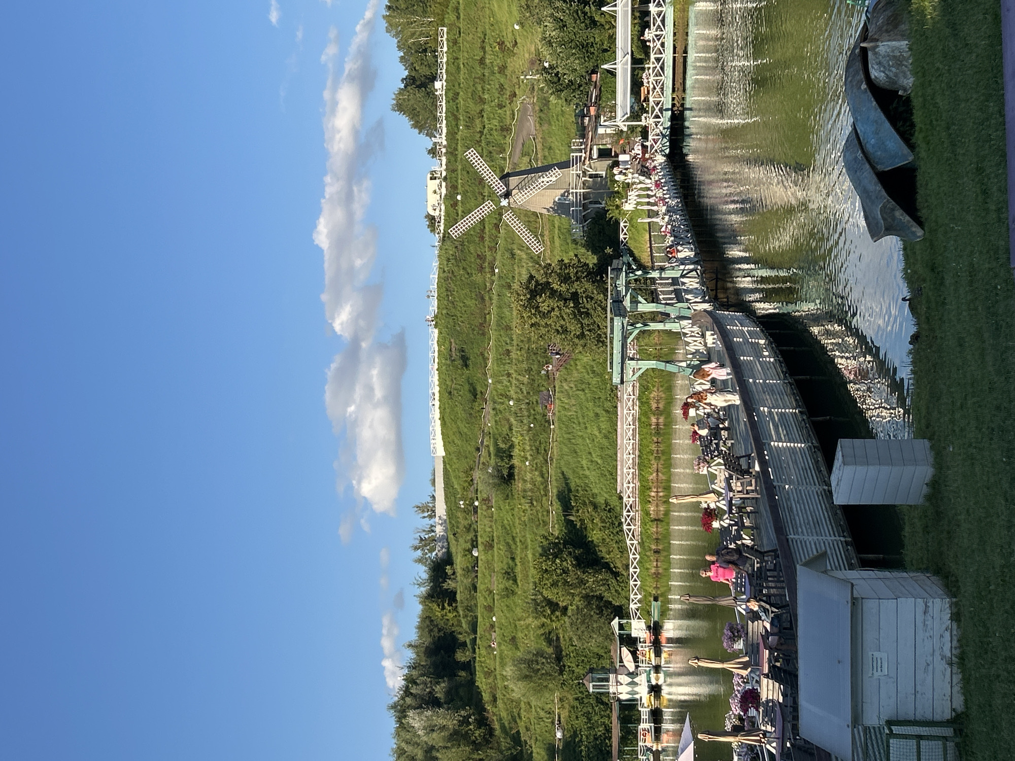 The image depicts a scenic view of a park area in Romashkovo, Russia, characterized by a tranquil lake, lush green hills, and architectural elements. A wooden boardwalk with tables and umbrellas, suggesting an outdoor dining area, stretches across the water. Several people are visible seated at tables, engaged in conversation or dining. Further back, a picturesque windmill stands on a hillside, adding a distinctive charm to the landscape. The hill is terraced and covered with green grass, with pathways winding up its slope. The sky above is a clear blue with scattered white clouds. The overall scene conveys a peaceful and idyllic atmosphere, typical of a recreational park.