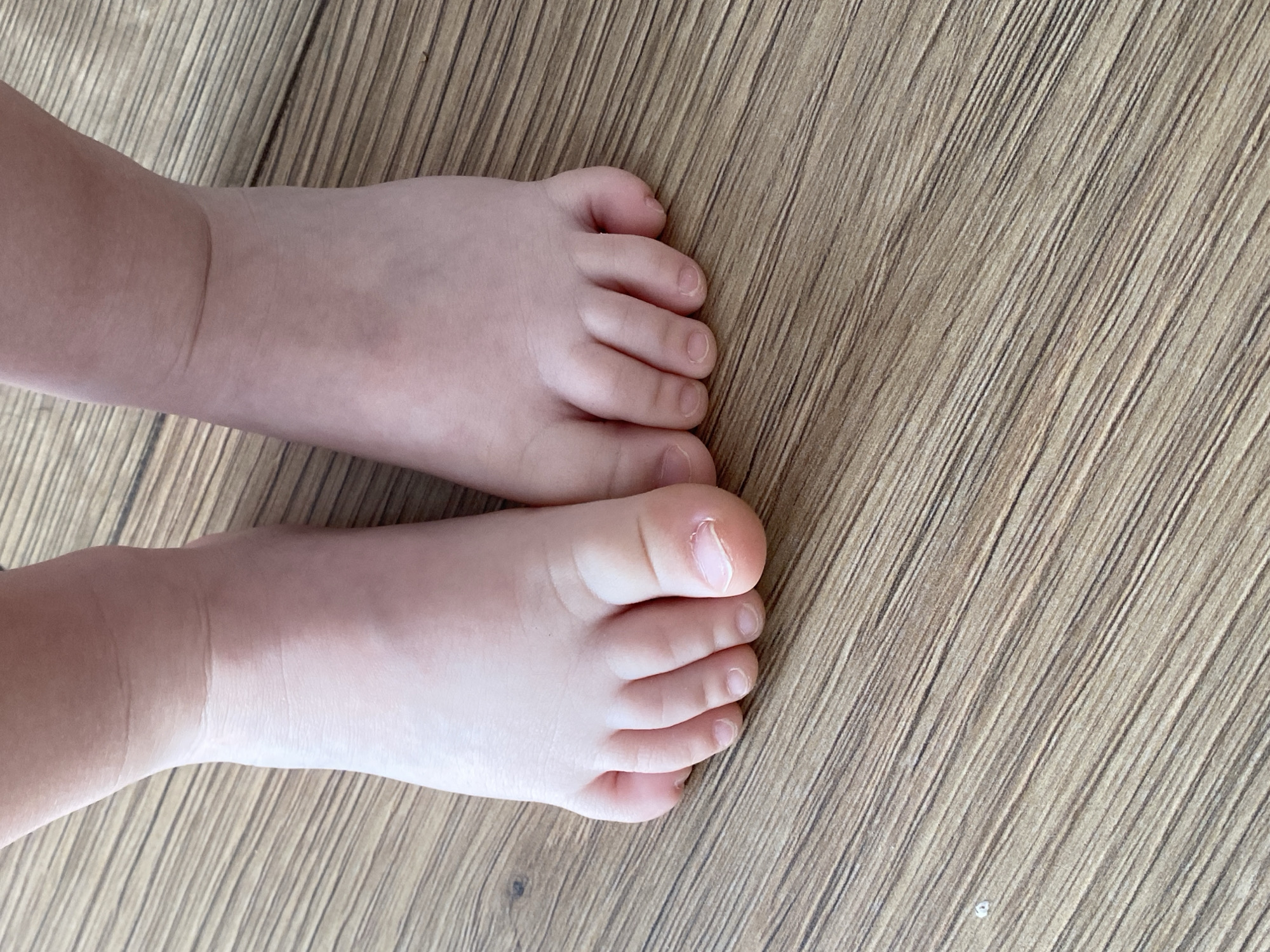 A close-up, high-angle shot shows two bare baby feet with pale skin resting on a light brown wooden floor with visible grain. The toes are plump and slightly curled, with visible toenails. The left foot's big toe is closest to the camera, and a small bit of dirt or lint is visible on the floor to the left of the feet. The lighting is bright and even, casting no significant shadows.