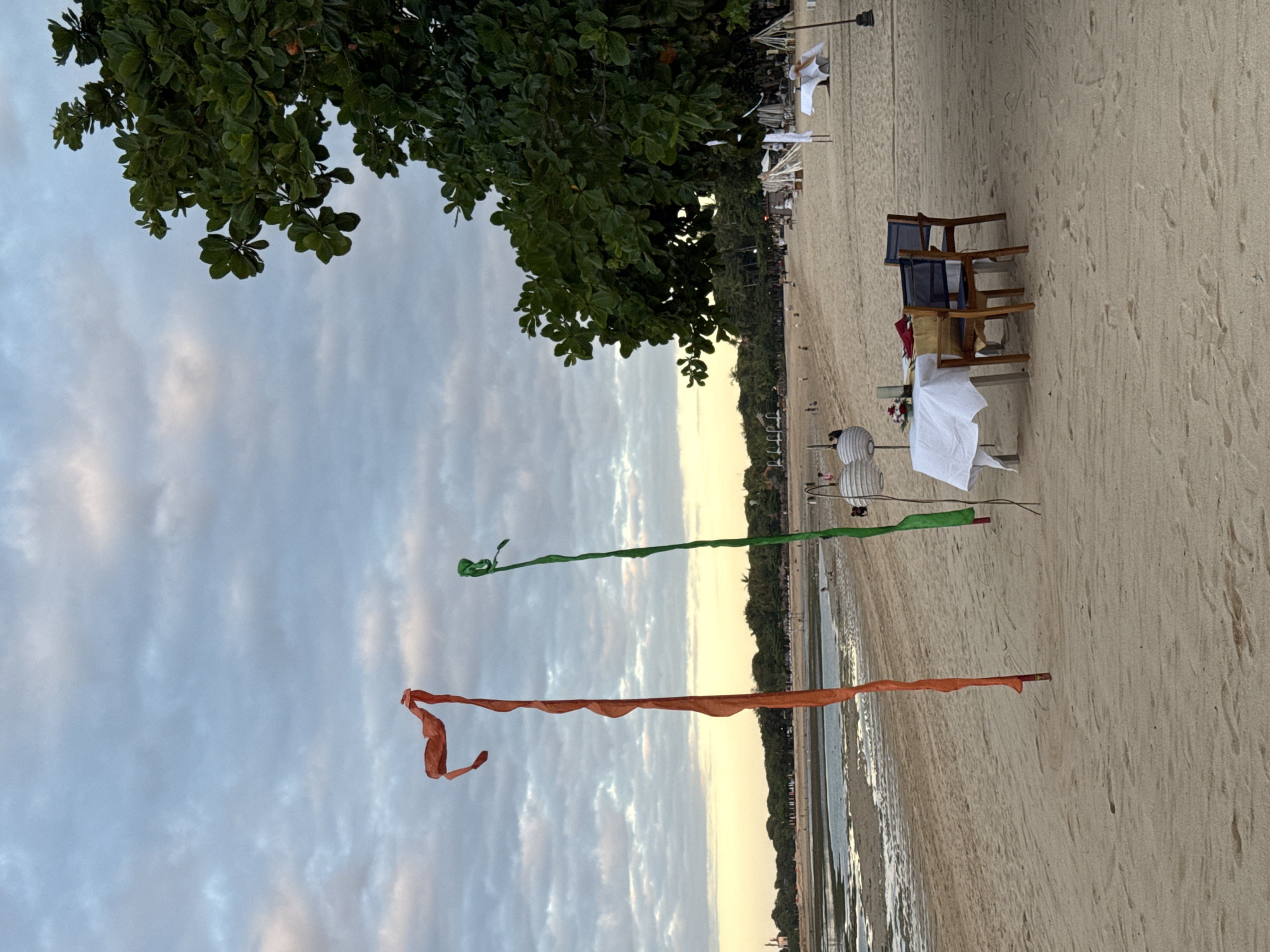 The image depicts a beach scene in Kuta Selatan, Indonesia. The foreground shows a sandy beach with two colored streamers, one green and one orange, attached to wooden posts, seemingly marking a path or area. Further back, a table with a white tablecloth is set up with chairs, potentially for a beachside dining experience. The background includes the ocean, the shoreline, and trees lining the coast. The sky is partly cloudy, suggesting either sunrise or sunset.