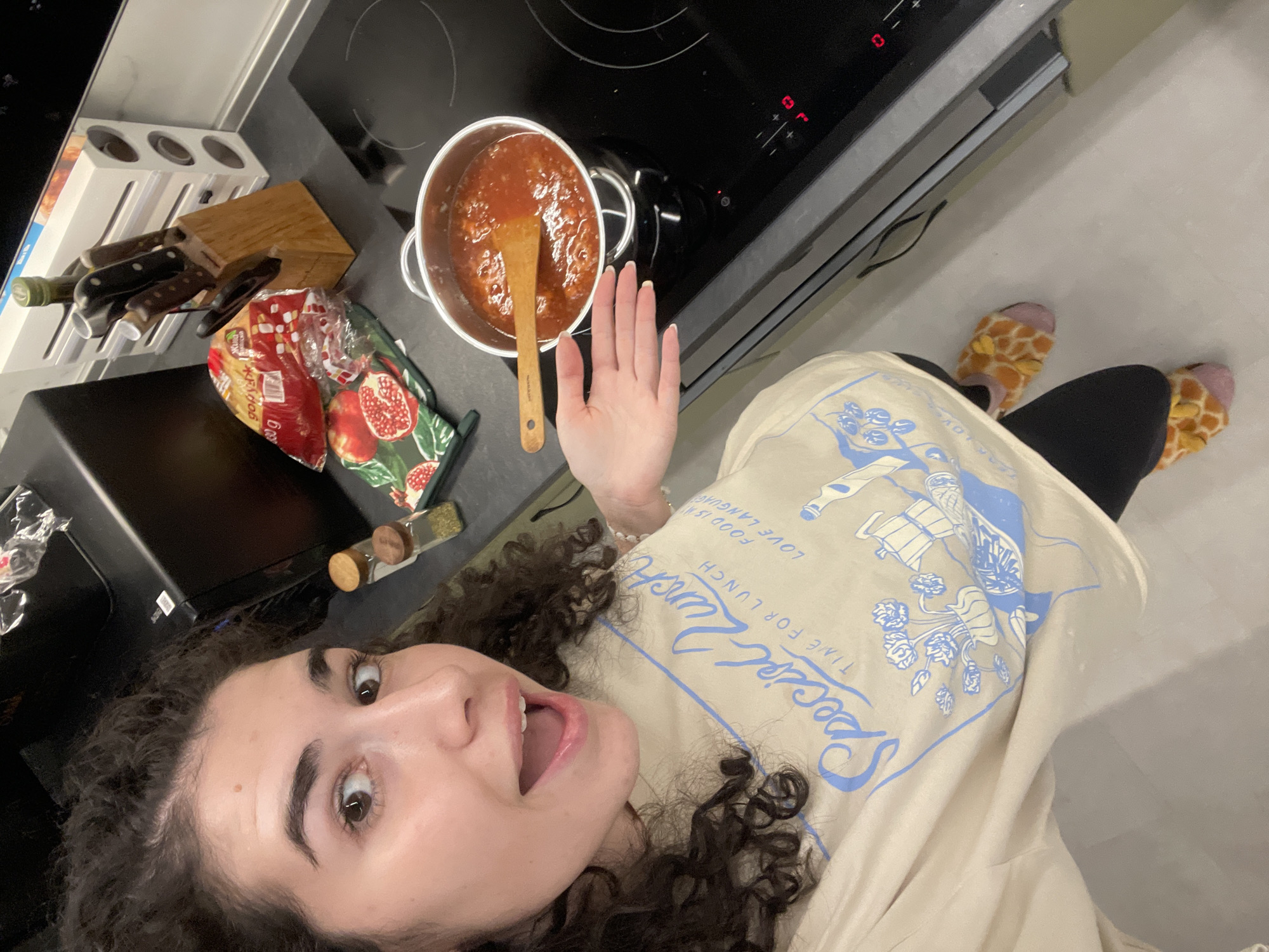 A woman with curly brown hair is taking a selfie in a kitchen. She is wearing a light beige shirt with blue designs and the words "Special Lunch, time for lunch, love food is my love language." She is also wearing black leggings and giraffe-patterned slippers. Her mouth is open in a surprised expression, and she is holding her hand up in a wave. A pot of tomato sauce is on the stovetop next to a wooden spoon. A knife block, a bag of pasta, and a pomegranate-patterned potholder are on the counter nearby. Two spice jars are on the left side of the pot. The city of Sofia, Bulgaria is not discernible in the image.