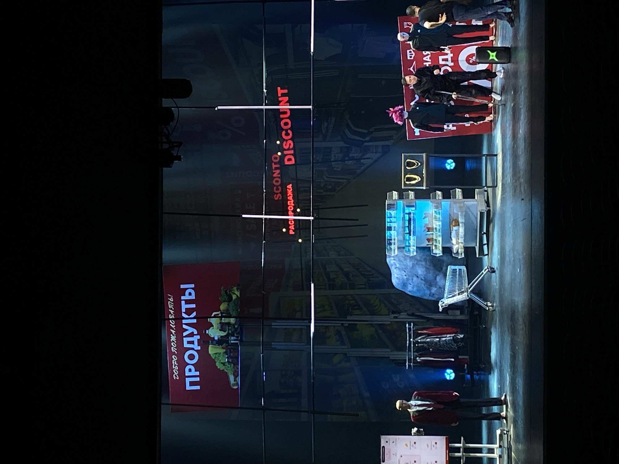 Here is a detailed description of the image:
The image captures a scene from what appears to be a stage performance or presentation, set in an unknown location. The backdrop consists of a large screen display, divided into multiple sections. On the left side of the screen, there is a display with the word "ПРОДУКТЫ" (Products) in large, bold lettering. Smaller text below it reads "ДОБРО ПОЖАЛОВАТЬ!" (Welcome!). The right side of the screen features the word "DISCOUNT" in prominent red letters, alongside smaller text in a different language that includes the words "SCONTO" and "РАСПРОДАЖА" (Sale).
The stage itself is a mixture of realism and abstraction. A few people are standing on the stage, including some wearing what appears to be masks or unusual headwear. They stand near large, red signs. In the center of the stage, there's a display resembling a grocery store section, complete with shelves, a shopping cart, and racks of clothing. Toward the front, a man stands beside a small sign, seemingly giving a presentation. The overall tone of the scene suggests a creative and possibly satirical take on the theme of shopping and discounts.
