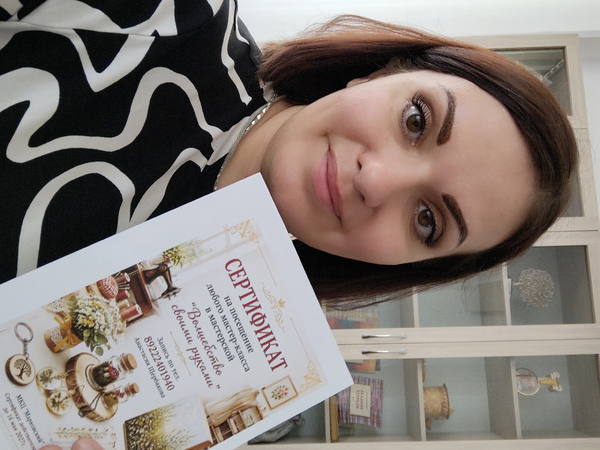 A woman with brown hair and hazel eyes holds up a certificate in a portrait format, tilting her head slightly. The certificate, written in Russian, is for a visit to a workshop called "Magic with Your Hands," and includes a phone number and name of a person named Anastasia Shcherbakova. The workshop is located in the "Markovskii" cultural center. The background consists of a cabinet with glass doors and various items displayed inside. The woman is wearing a black and white patterned top and a silver chain around her neck.