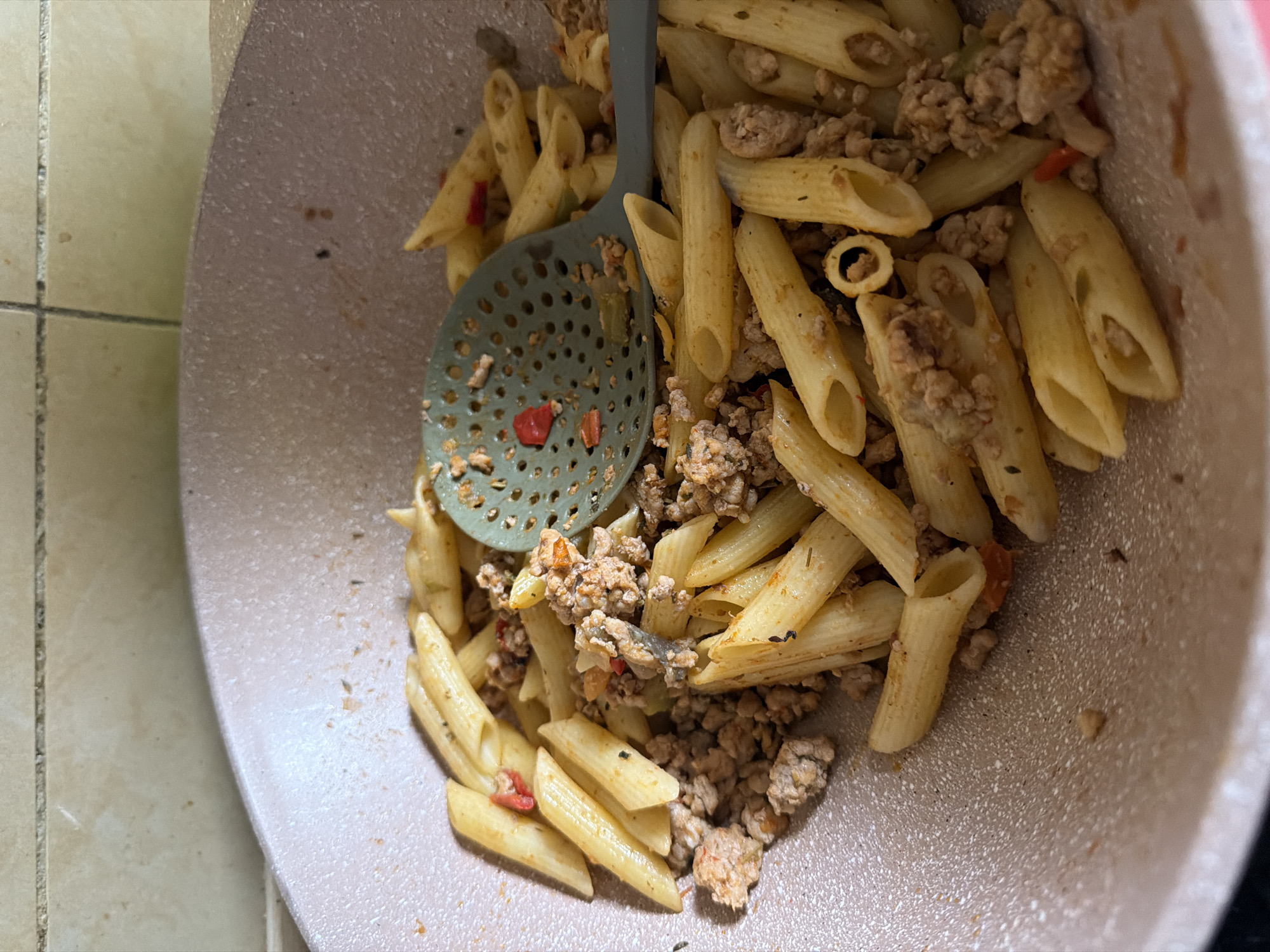 The image shows a close-up, overhead view of a pan containing pasta with ground meat and some red pepper pieces. A green slotted spatula is partially submerged in the pasta, with some of the red pepper stuck to it. The pasta appears to be penne. The pan has a speckled, light pinkish-beige interior and is set against a backdrop of beige tiled walls. There is no visible indication of the city of Figueres, Spain.