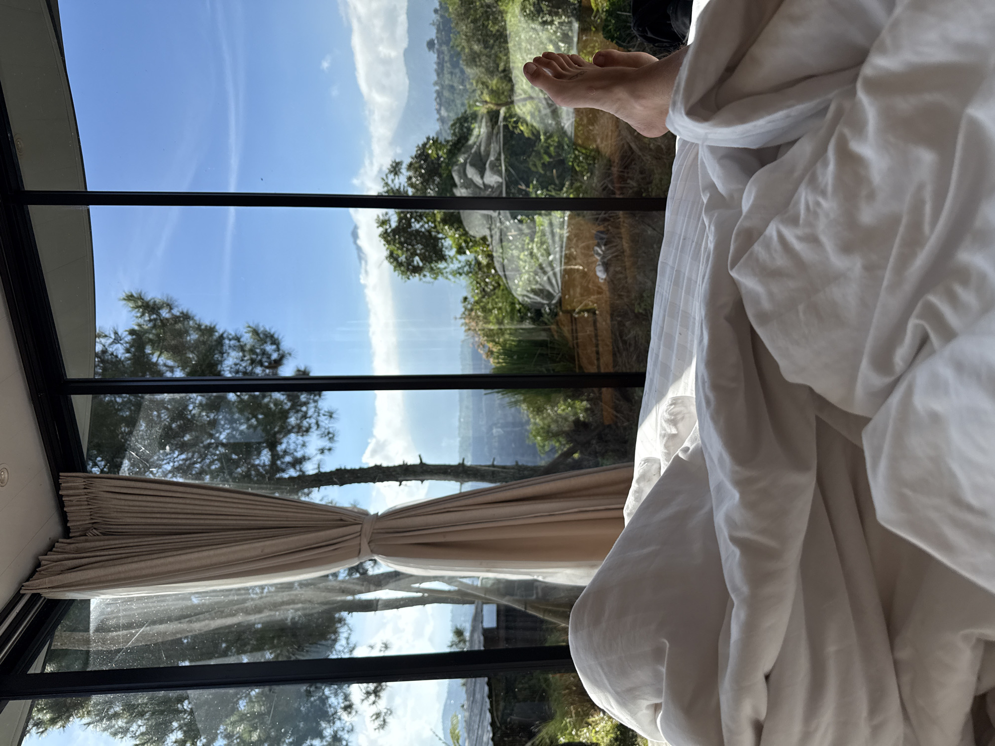 The image shows a view from inside a room in Kintamani, Indonesia. The viewer is likely in bed, evidenced by the white, rumpled bedsheets dominating the right side of the frame and a pair of bare feet visible at the top. The room features large, multi-pane windows with black frames that offer a panoramic view of the surrounding landscape. A beige curtain is tied back, partially obstructing the view. The exterior scene includes lush green vegetation, trees, hills, and a clear blue sky with scattered clouds. The composition suggests a peaceful, tranquil setting in a natural environment.