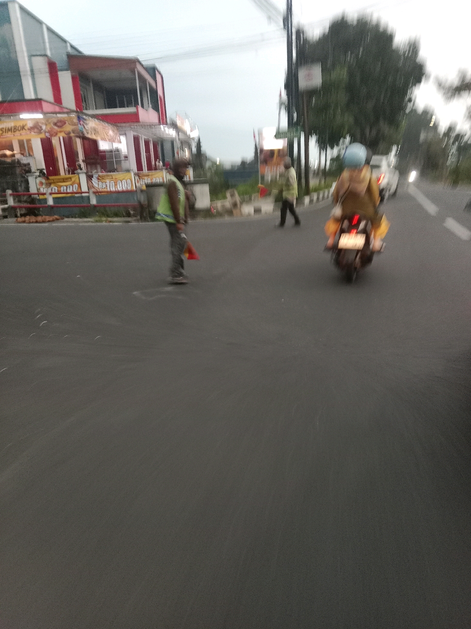 The image depicts a street scene in Kalianget, Indonesia, during overcast daylight. In the foreground, a person in a high-visibility vest and dark pants is walking on the asphalt road, carrying a red flag in their right hand. Behind them, further down the road, a person is riding a motorcycle, also in motion, with bags strapped to the back. To the left of the road, a building with red and white accents is visible, with signage indicating "SIMBOK" and what appears to be a food stall or restaurant. The overall impression is one of a moderately busy street, possibly on the outskirts of a commercial area, with ongoing pedestrian and vehicular traffic.