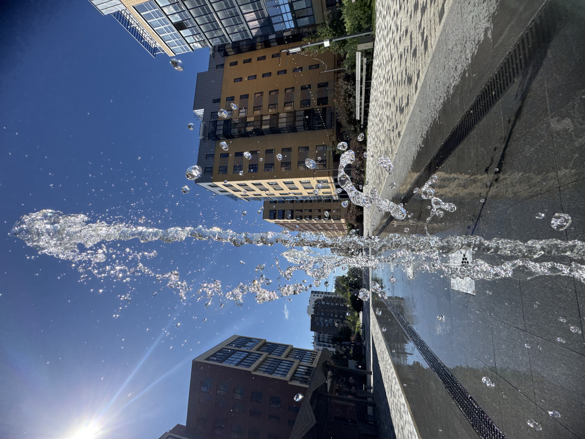 This is a close-up, low-angle shot of a fountain jet on a sunny day in the city of Novoselye, Russia. The water jet is centrally located and appears to be a single, powerful stream, with many droplets suspended in the air around it. In the background, modern, multi-story apartment buildings with dark windows and balconies are visible. The ground is paved with light-colored stones, and there are dark, linear drains running parallel to the fountain. The sky is a clear, bright blue, with the sun creating lens flare to the left of the frame. The environment appears to be an urban public space, likely a courtyard or plaza within a residential complex. There are no people or animals visible in the frame.