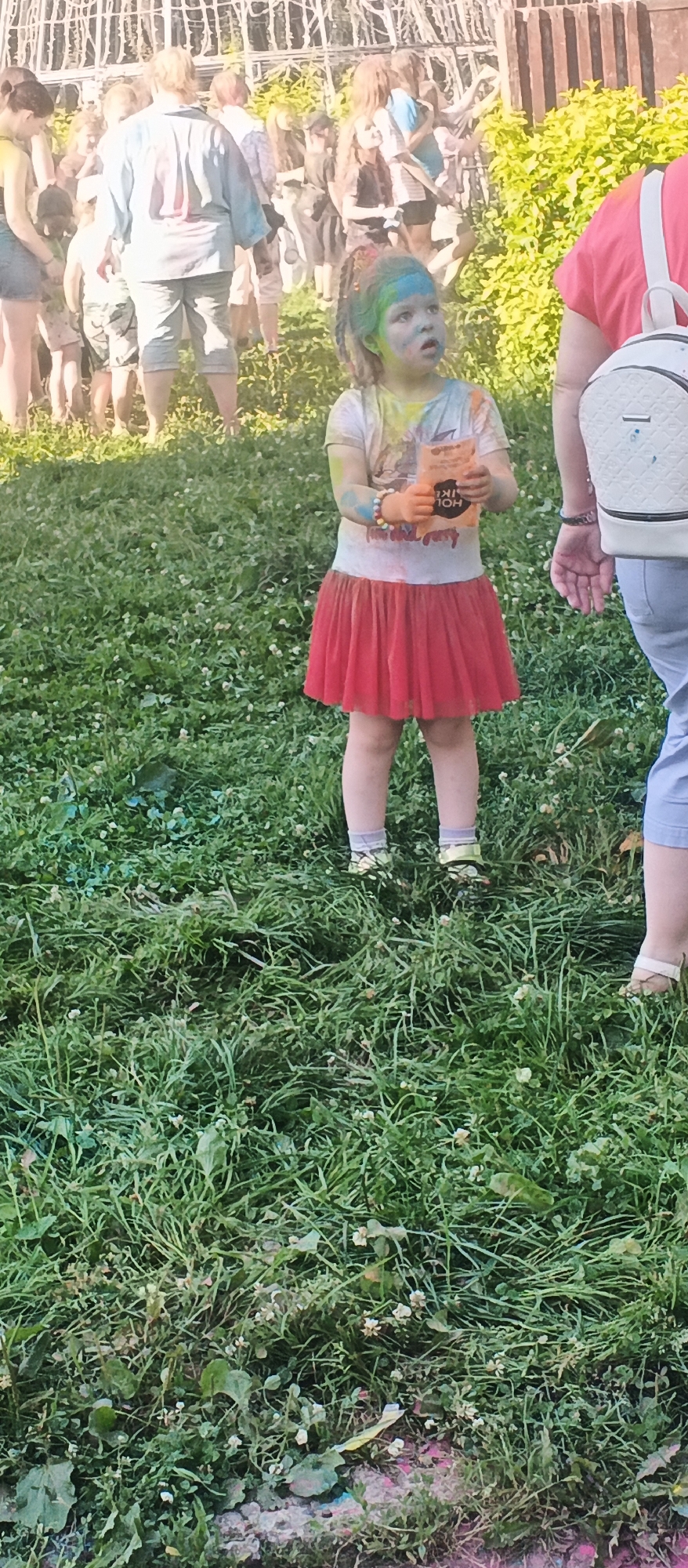 A young girl with rainbow-colored streaks in her hair, wearing a white t-shirt and a red skirt, stands in a grassy area. Her face and t-shirt are smudged with colorful powder, suggesting participation in a festival or event like Holi. She holds a small orange packet in her hands. Behind her, a crowd of people of various ages are visible, many also appearing to have powder on them. The background includes a metal fence-like structure and green foliage. The scene appears to be an outdoor gathering, possibly in a park or public space in Izhevsk, Russia, given the context provided.