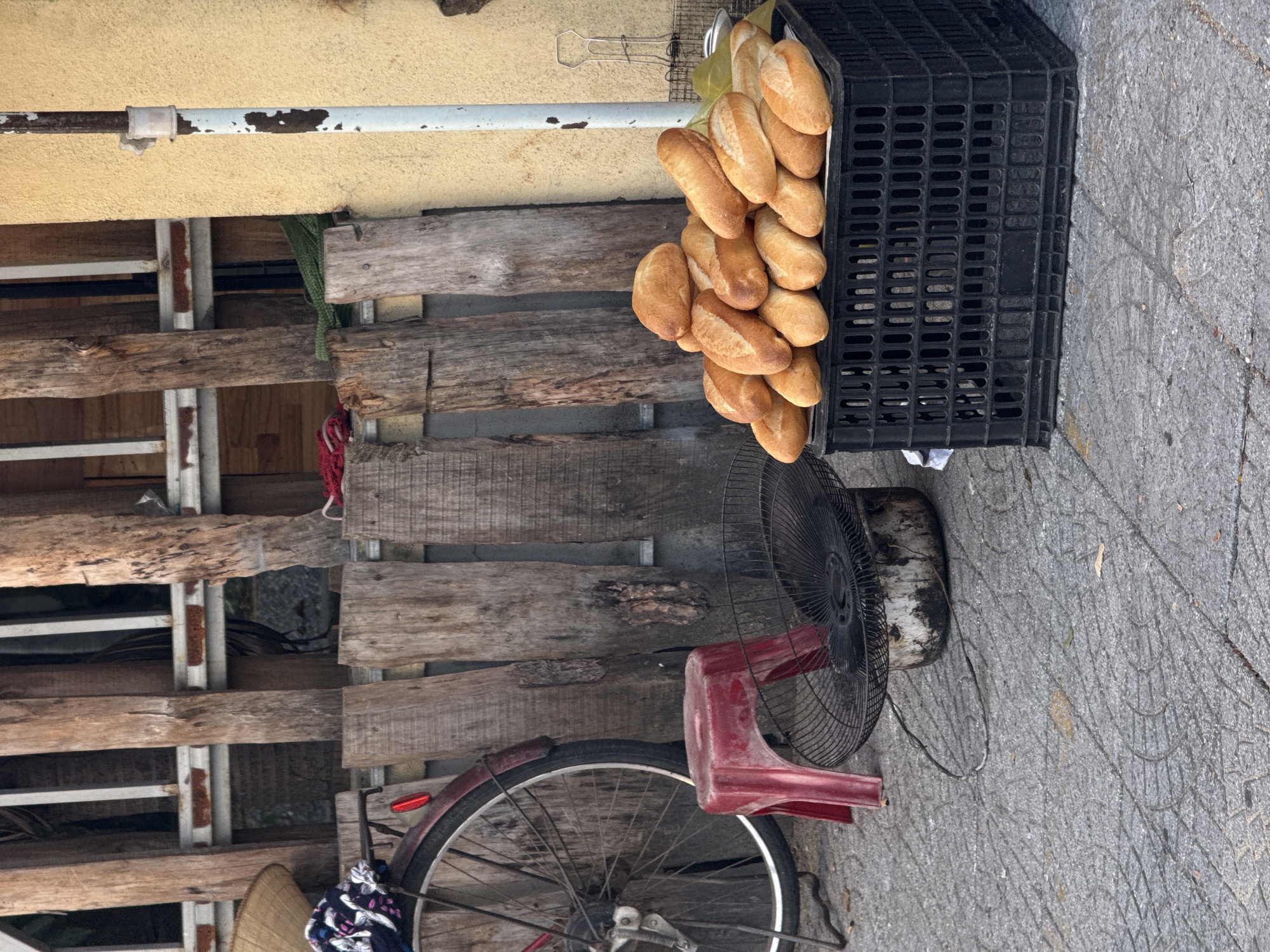 This is a street scene in Da Nang, Vietnam, featuring a collection of items arranged outdoors. In the foreground, a large black plastic crate overflows with golden-brown baguettes. To the left of the crate, a small red plastic stool sits next to an old, black electric fan with a circular grill, its base appearing stained and worn. Behind these items, a bicycle wheel is visible, its red fender and spokes suggesting it's an older model. A straw hat is perched on top of a colorful cloth draped over the bicycle's rear. The background consists of a weathered wooden fence or wall, constructed from vertical planks. A yellow stucco wall and a rusted metal pole are visible to the right of the wooden structure. The ground is paved with grey interlocking concrete tiles. The overall impression is of a simple, utilitarian outdoor setup, possibly associated with a street vendor or a household.