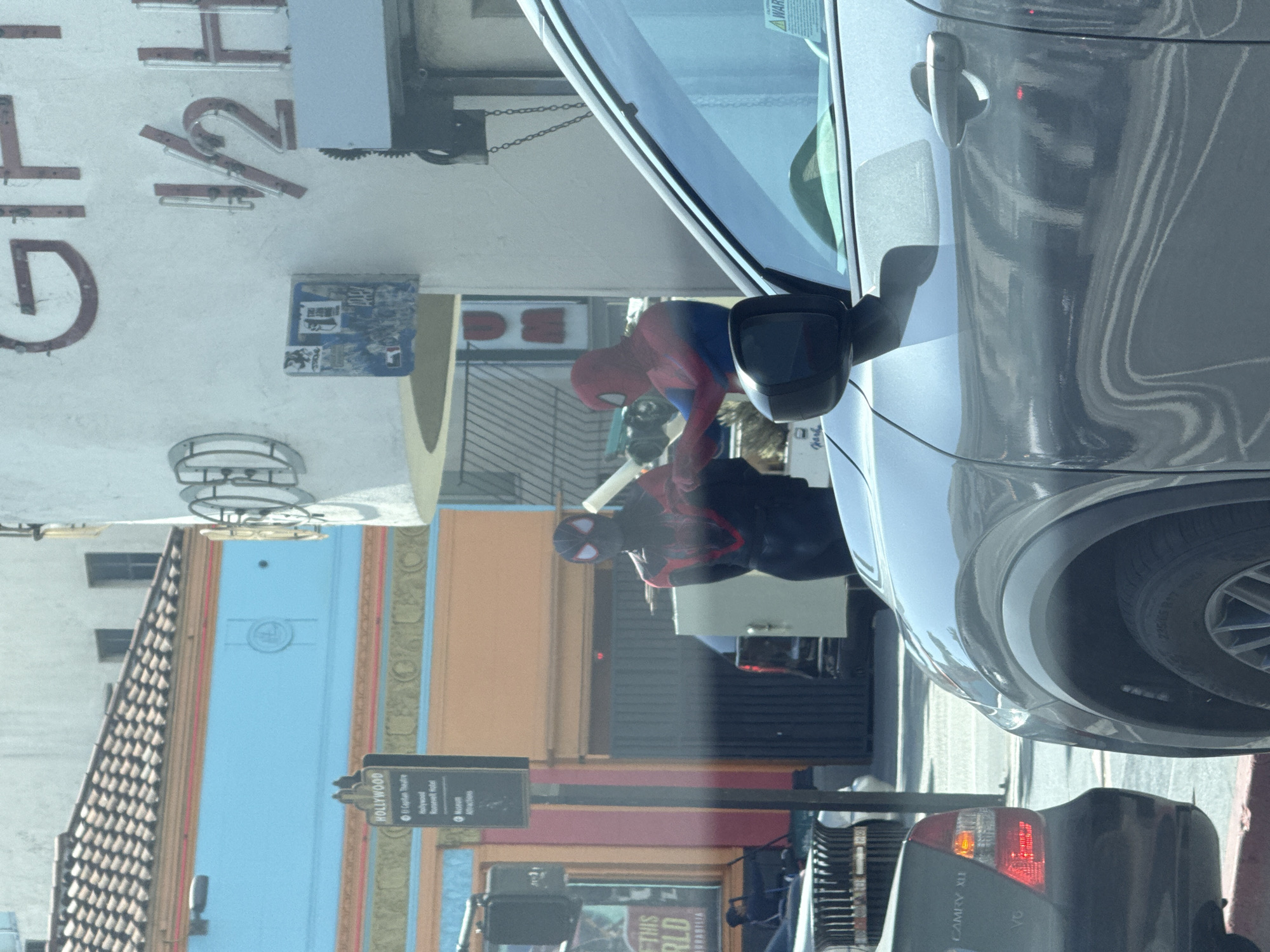 This image captures a vibrant street scene in Hollywood, Los Angeles, during daytime under clear skies. The perspective is from the interior of a gray car, looking outwards.
The primary subjects are two individuals dressed as Spider-Man characters. One is wearing the classic red and blue suit, while the other is in a black and red suit, reminiscent of Miles Morales. They appear to be standing or walking on the sidewalk or street, with one character slightly more in the foreground than the other.
The setting is an urban street lined with multi-story buildings, featuring facades in light blue, orange, and white. A prominent black street sign in the mid-ground helps identify the location, displaying "HOLLYWOOD" at the top, followed by bullet points for "El Capitan Theatre," "Hollywood Roosevelt Hotel," and "Attractions." Another building features large, red, partially obscured letters that read "GIFT 1/2 H."
In the foreground, the side and front of the gray car from which the photo is taken are visible, with reflections on its surface. Behind the Spider-Man characters, another dark gray car is visible on the street, identifiable by the text "CAMRY XLE" and "V6" on its rear bumper, with its brake lights illuminated. A poster or sign on a building in the background has partially visible text "THIS RLD." The scene suggests a busy tourist area, typical of Hollywood Boulevard, where costumed performers often engage with visitors.