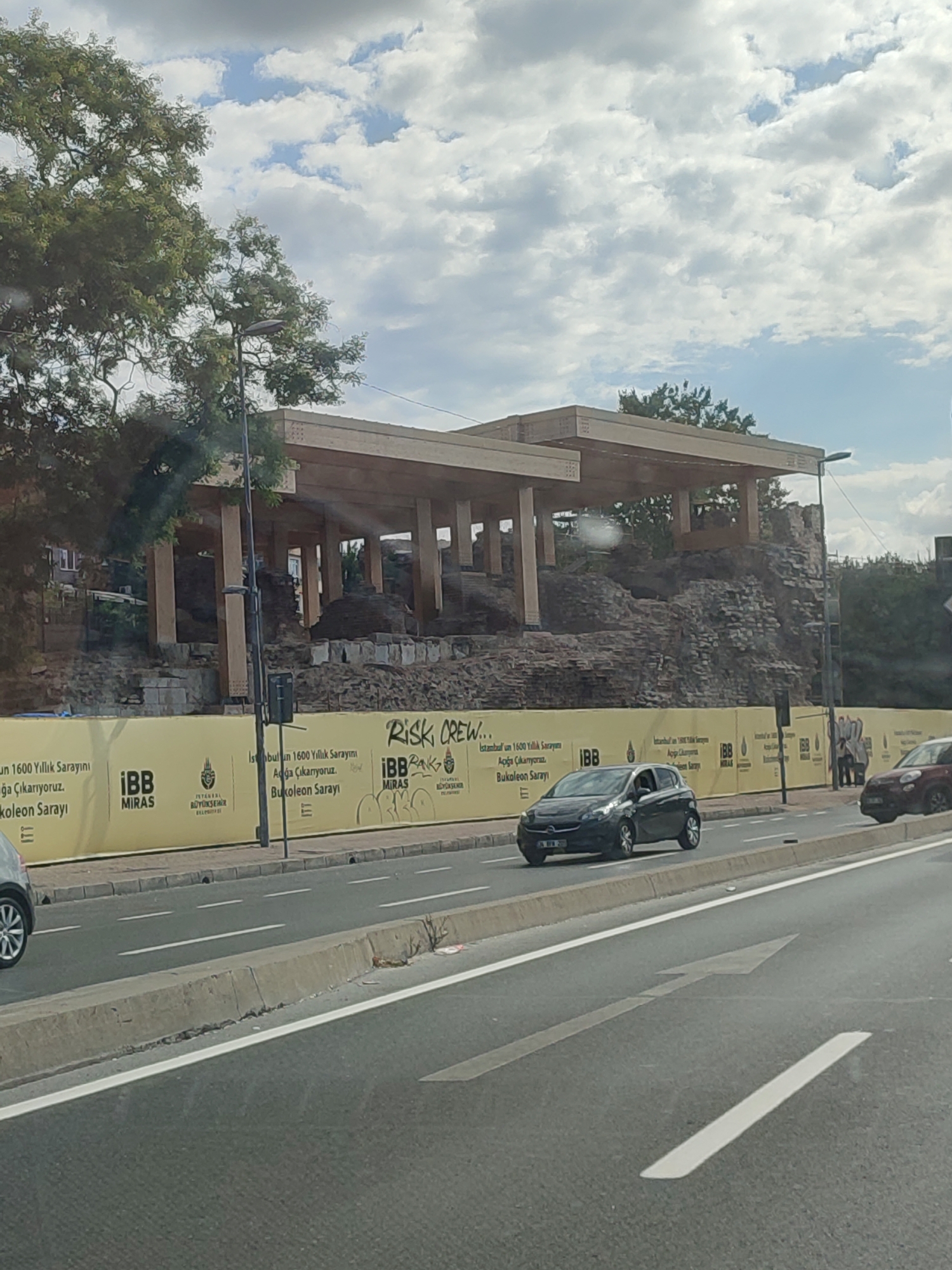 The image shows an outdoor scene in Istanbul, Turkey, featuring a road with passing vehicles and an archaeological site under construction in the background. The site is cordoned off with a yellow banner displaying Turkish text and logos, including "IBB MIRAS" and the Istanbul Metropolitan Municipality emblem. Above the banner, the ruins of an old structure are visible, with a modern wooden roof structure erected over it. A street light and trees are also present. The road has lane markings and a concrete divider. Two cars are on the road, one dark-colored in the foreground and a red one further back. The sky is partly cloudy.