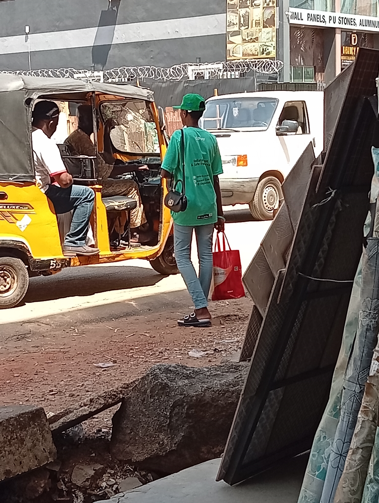 The image depicts a street scene in Maiburiji, Nigeria. A yellow "keke" (auto rickshaw) is partially visible on the left side of the frame, with two male passengers and a driver. A person wearing a green shirt, jeans, and a green hat stands on the street holding a red shopping bag and a black purse. A white van is parked behind the auto rickshaw. The background includes buildings with signs advertising "WALL PANELS, PU STONES, ALUMINUM." Construction materials are seen in the foreground, partially obscuring the view of the street. The street surface appears uneven and possibly under construction.