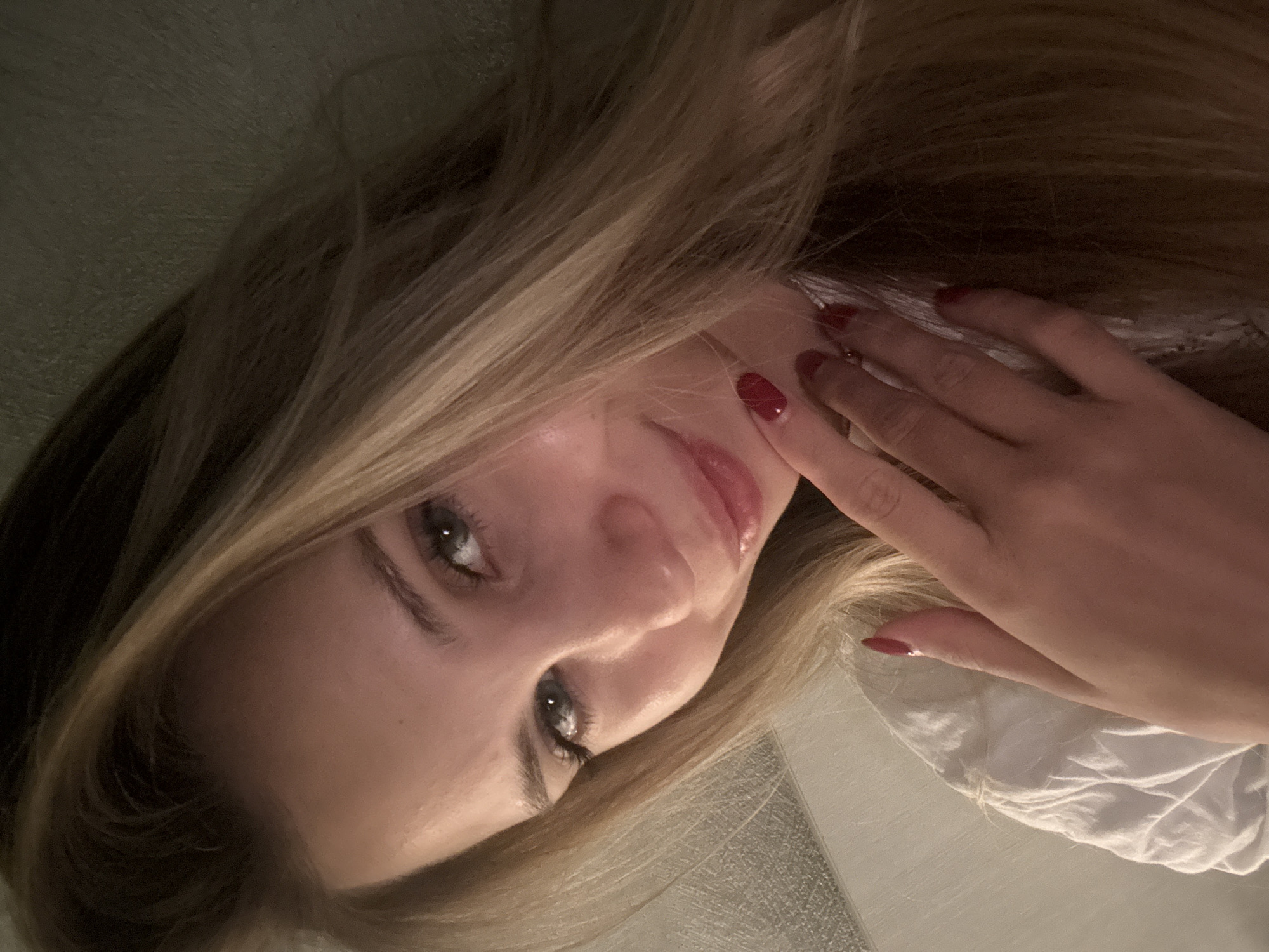 The image shows a close-up, head-and-shoulders portrait of a young woman with long, blonde hair. She is lying down, with her head tilted and her face angled towards the viewer. Her eyes are open and looking directly at the camera. She has makeup on, including mascara and what appears to be lip gloss, which gives her lips a shiny appearance. Her right hand is resting on her cheek, and her fingernails are painted red. The background is out of focus but appears to be a textured surface, possibly a wall or bedding, with some hints of light-colored fabric. The lighting is soft and appears to be coming from the side, creating highlights on her face and hair. The overall mood of the image is intimate and slightly melancholic. There is no visible text in the image. The setting appears to be indoors, possibly a bedroom, with no specific cues indicating the time of day or weather.