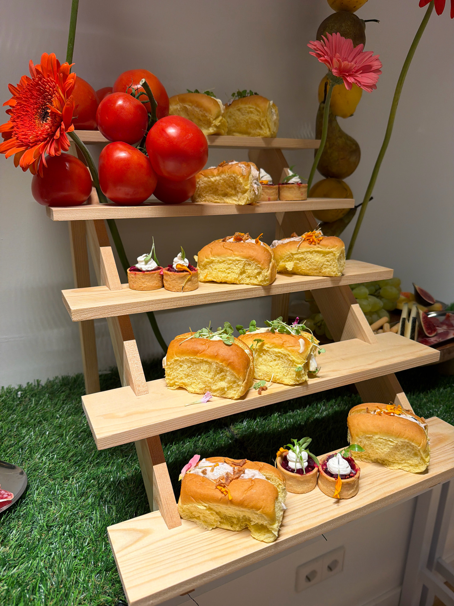 A tiered wooden display stand, resembling a miniature picnic table, is set up on a patch of artificial grass. The stand is laden with various food items and decorative elements. The top shelf holds several bright red tomatoes and an orange gerbera daisy. Below that, on the second shelf, are small round pastries filled with a dark red substance and topped with white cream and green sprouts, alongside some yellow bread rolls. The third shelf features more yellow bread rolls, some topped with microgreens and garnishes. The bottom shelf also displays yellow bread rolls. In the background, to the right, are bunches of green grapes, figs, and what appears to be cured meat and cheese. The overall setting suggests a catered event or a styled display. The location is identified as Ibiza, Spain.