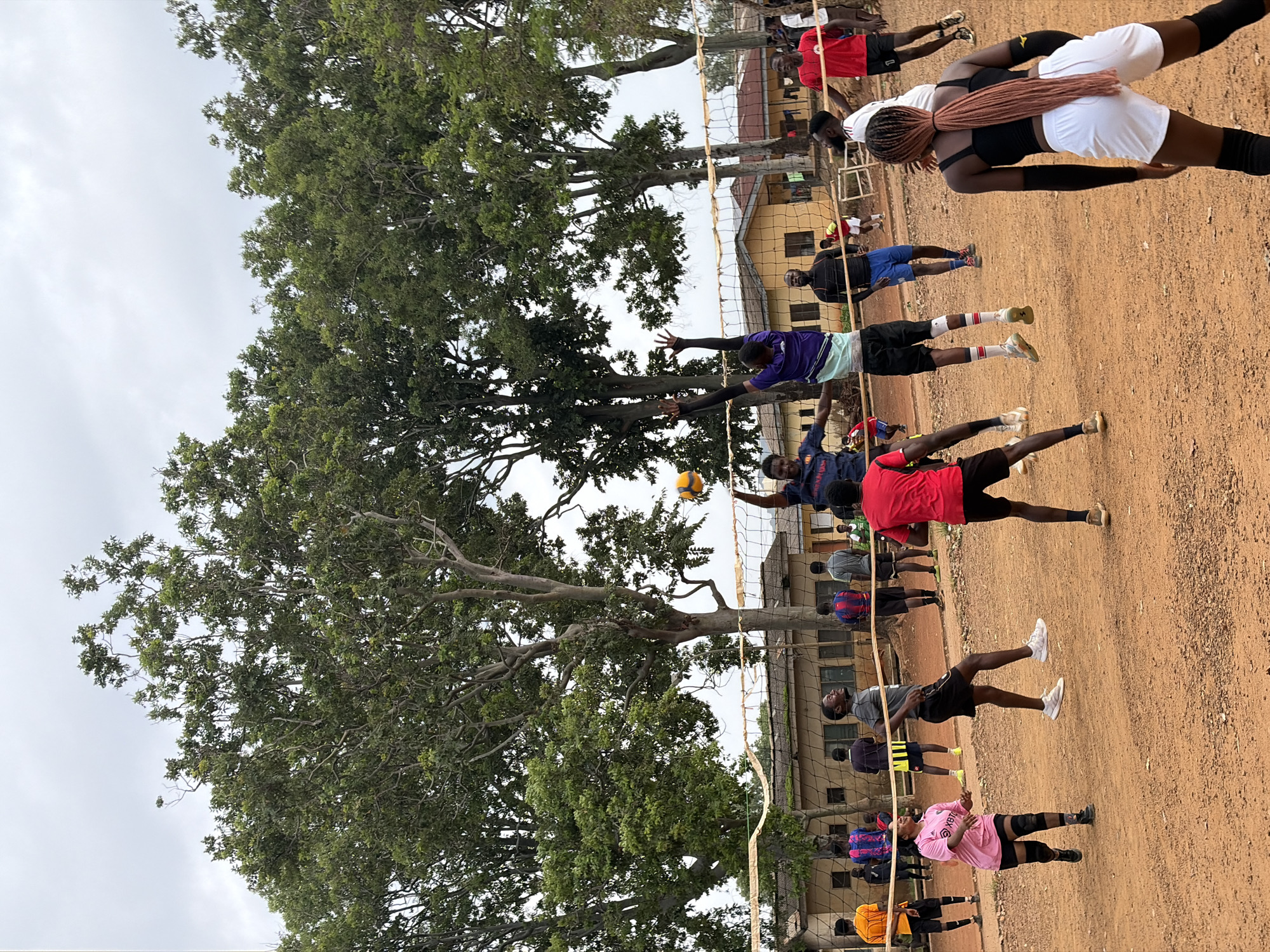In Rafin Par, Nigeria, a group of people are engaged in a volleyball game on a dirt court. The game appears to be in progress, with several players in mid-action. In the foreground, a woman with braided brown hair, wearing a black tank top and white shorts, is facing away from the camera, with her arms bent at the elbows, positioned as if to receive a volleyball. To her left, a man in a pink shirt and black shorts, also with his arms extended, is looking up. Several other players are on the court, some in the process of hitting or blocking the ball, while others are observing the game. A volleyball net is stretched across the court. In the background, there are trees with lush green foliage and several buildings with yellowish walls and brown roofs. The sky is overcast with a grayish hue. The ground is predominantly reddish-brown dirt, typical of an outdoor playing surface. The scene captures a moment of active recreation within a community setting.