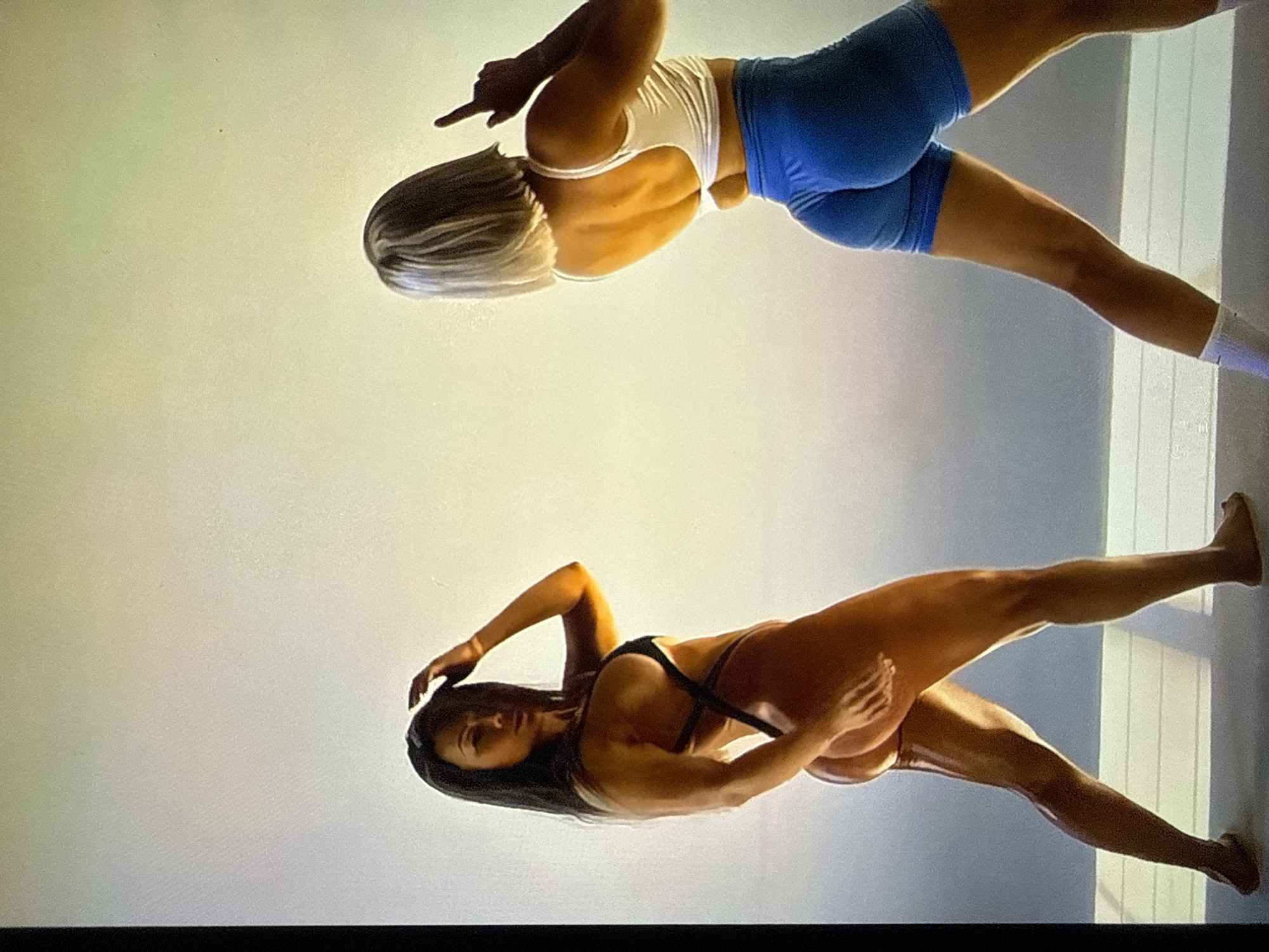 The image depicts two female individuals posing in what appears to be a studio setting with a plain, light-colored background. The location is Unknown, Unknown.
The individual on the left stands barefoot, angled slightly to her right, with her head turned towards the viewer. She has long, dark hair and wears a black, strappy one-piece garment that exposes parts of her back and glutes. Her right hand is raised to touch her head, and her left hand rests on her left thigh. She exhibits a muscular physique with visible highlights on her skin, suggesting a reflective surface or oil.
The individual on the right stands with her back mostly facing the viewer, angled slightly to her left. She has short, light-colored (blonde) bob-length hair. She wears a white sports bra and form-fitting blue athletic shorts. Her right arm is raised, with her index finger pointing upwards. Her left foot is partially visible with a white sock. She also displays a muscular physique, with prominent glutes in the blue shorts.
The background is a uniform light gray or off-white color, suggesting a seamless backdrop. A horizontal line is visible across the lower portion of the image, indicating a floor-to-wall transition or a platform edge. The lighting is bright and diffuse, creating subtle shadows beneath the individuals. A dark vertical bar is present along the far left edge of the image.