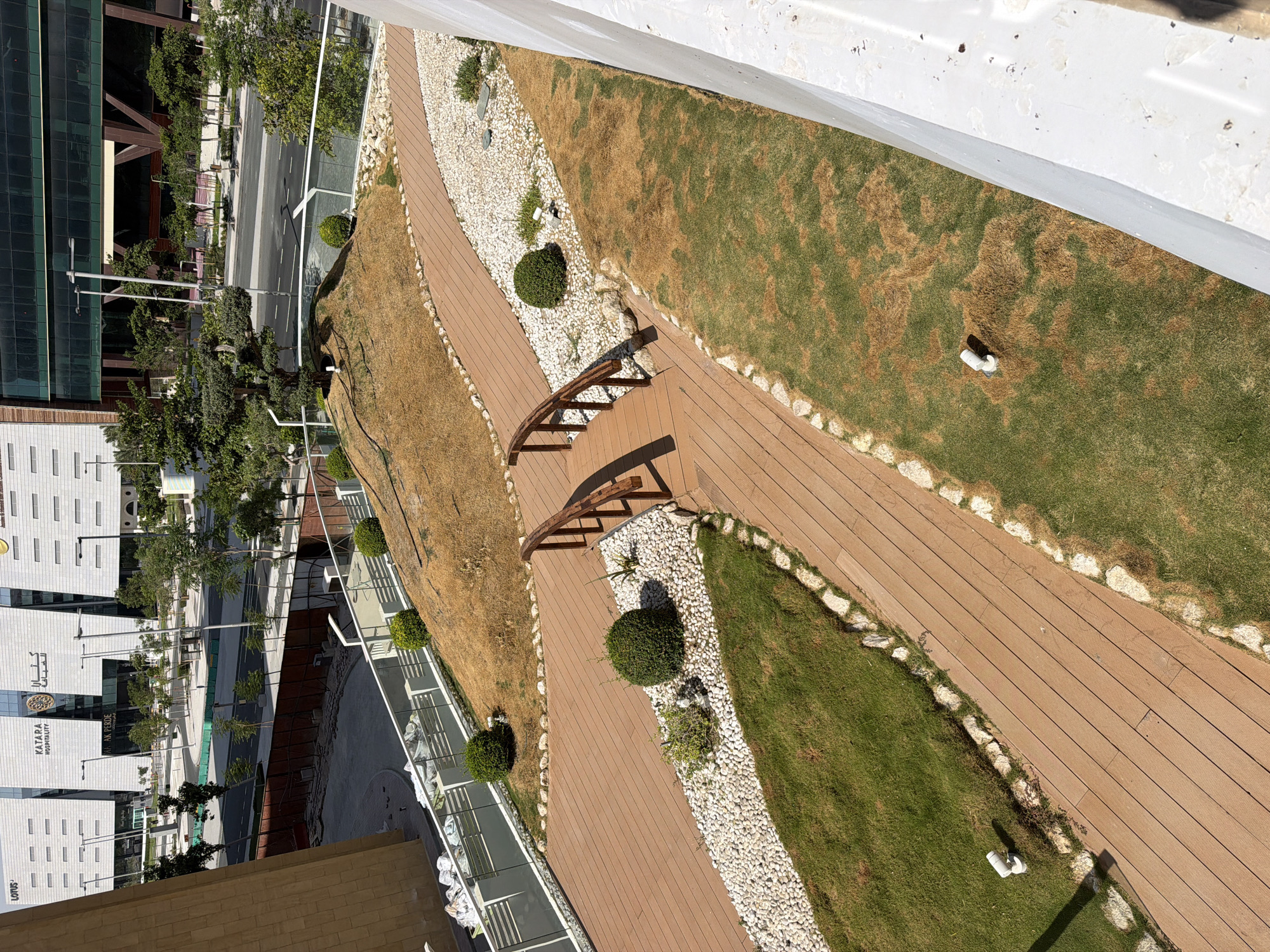 An aerial, high-angle, outdoor, daytime, medium shot captures a modern urban landscape with a focus on a tiered, landscaped garden area. The garden features wooden pathways, white pebble ground cover, patches of green grass, and several meticulously shaped green bushes. A small, arched wooden bridge crosses one of the pathways. In the background, sleek, contemporary buildings with glass facades dominate the skyline, interspersed with trees and streetlights. A road is visible in the middle ground, with some construction fencing in the foreground. The overall impression is one of an upscale, well-maintained urban development.