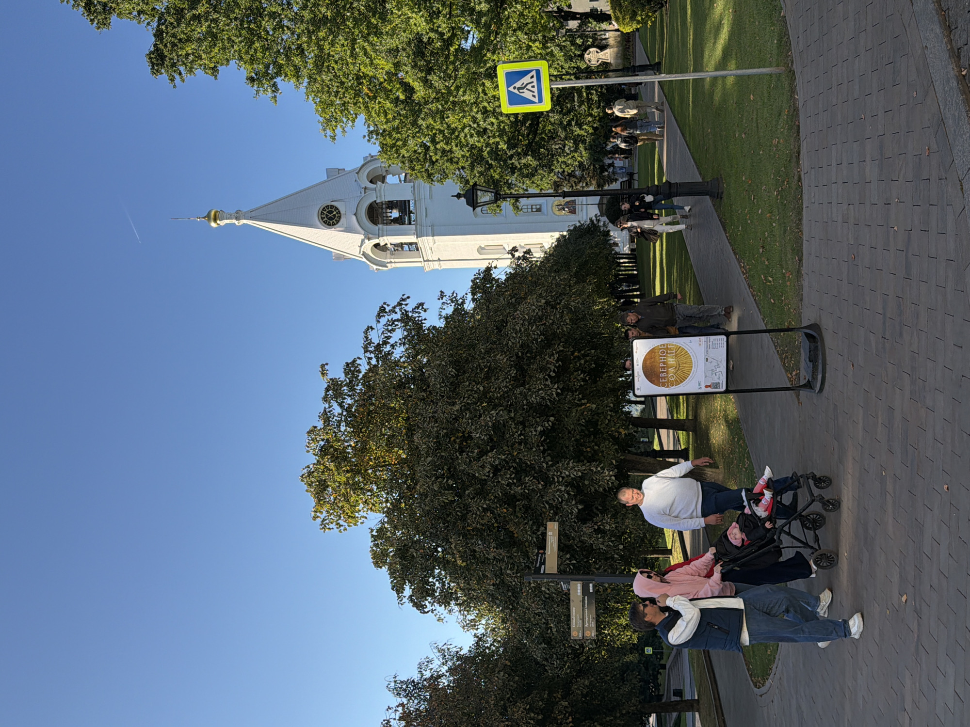 In Nizhny Novgorod, Russia, a bright, sunny day illuminates a scene in a park-like setting with a prominent white church tower in the background. A group of people are walking on a paved path. In the foreground, a man in a blue vest and jeans, a woman in a pink hoodie, and another man in a white sweatshirt are walking. The woman is pushing a stroller with a baby inside. Further back, other pedestrians are visible on the path and a lamppost stands near the church. A yellow pedestrian crossing sign is positioned to the right of the path. The church tower, with a golden dome and a clock face, stands tall against a clear blue sky. Trees with green foliage line the path and surround the tower. A sign with Cyrillic text is displayed on an easel near the foreground. The ground is paved with interlocking bricks, and patches of green grass are visible.