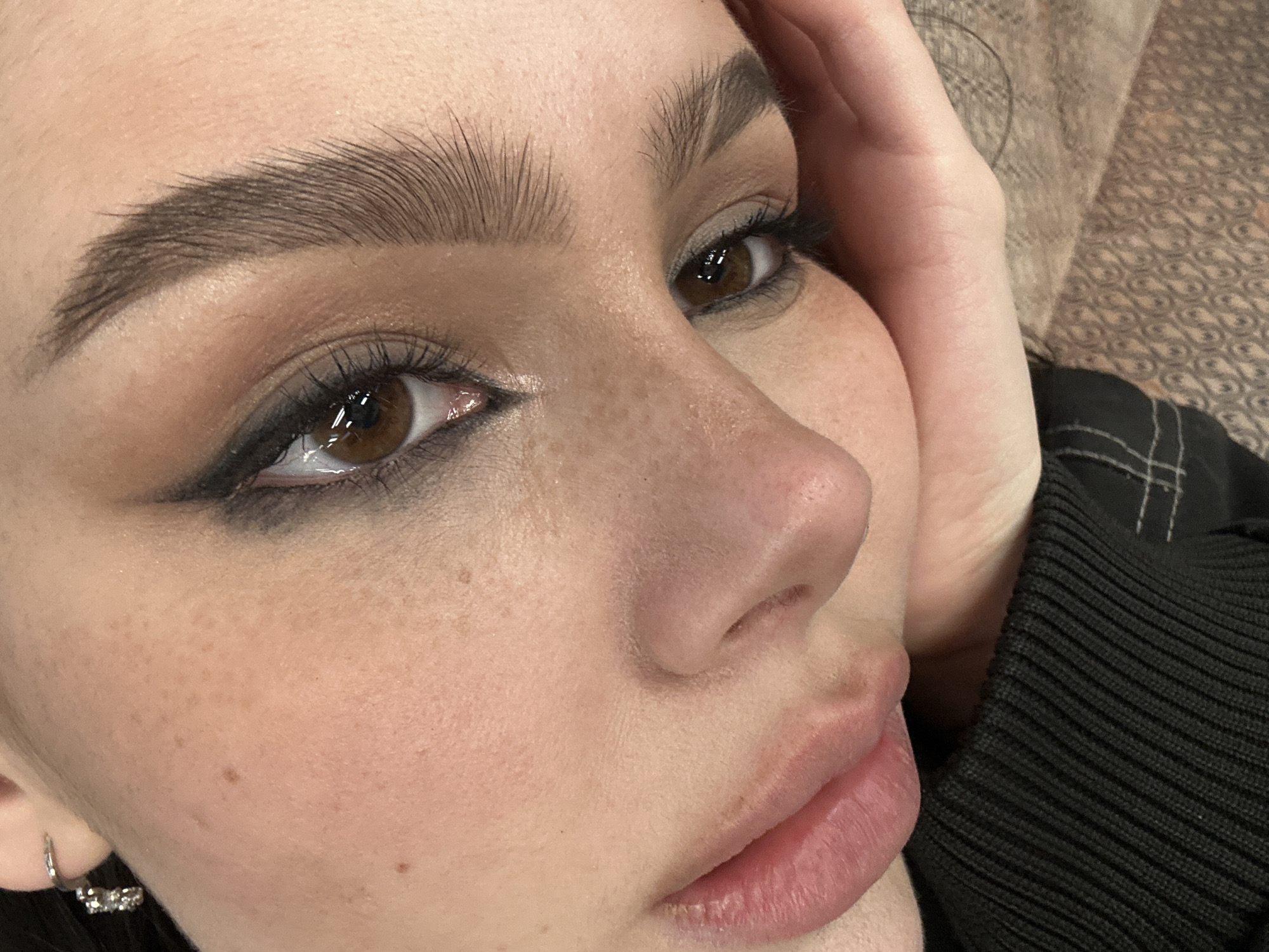 A close-up image shows a person's face with brown eyes, arched eyebrows, and fair skin with freckles. The person has dark eye makeup, including eyeliner and eyeshadow. Their lips are full and pinkish. Part of a hand is visible near the face, and a black garment is also in view. The background is slightly out of focus and appears to be a textured surface. There is no indication of a specific location, especially Moscow, Russia.