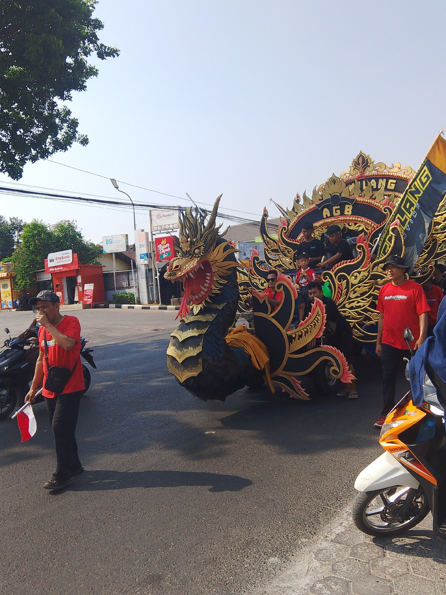 In the city of Rejosari, Indonesia, a parade is taking place on a street under a clear sky. The main subject is a large, ornate float shaped like a black and gold dragon. The dragon's head is detailed with red accents and its mouth is open. The float is decorated with gold trim and features the letters "AGB" prominently. Several individuals are visible on the float, including men and a young boy.
To the left of the float, a man wearing a red t-shirt, black pants, and a baseball cap walks while holding a small Indonesian flag. He has a black bag slung across his body. Behind him, several motorcycles are parked.
To the right of the float, another man in a red t-shirt and black pants stands near a motorcycle. A blue jacket is visible on a person partially in the frame to the far right.
The background includes buildings that appear to be shops or businesses, some with signage such as "Enduro," "BSI ATM," and "Ayam GIP." Overhead, power lines and a street lamp are visible. The street surface is asphalt with some hexagonal paving stones visible on the right side. The overall scene suggests a celebratory event or festival in an urban setting.