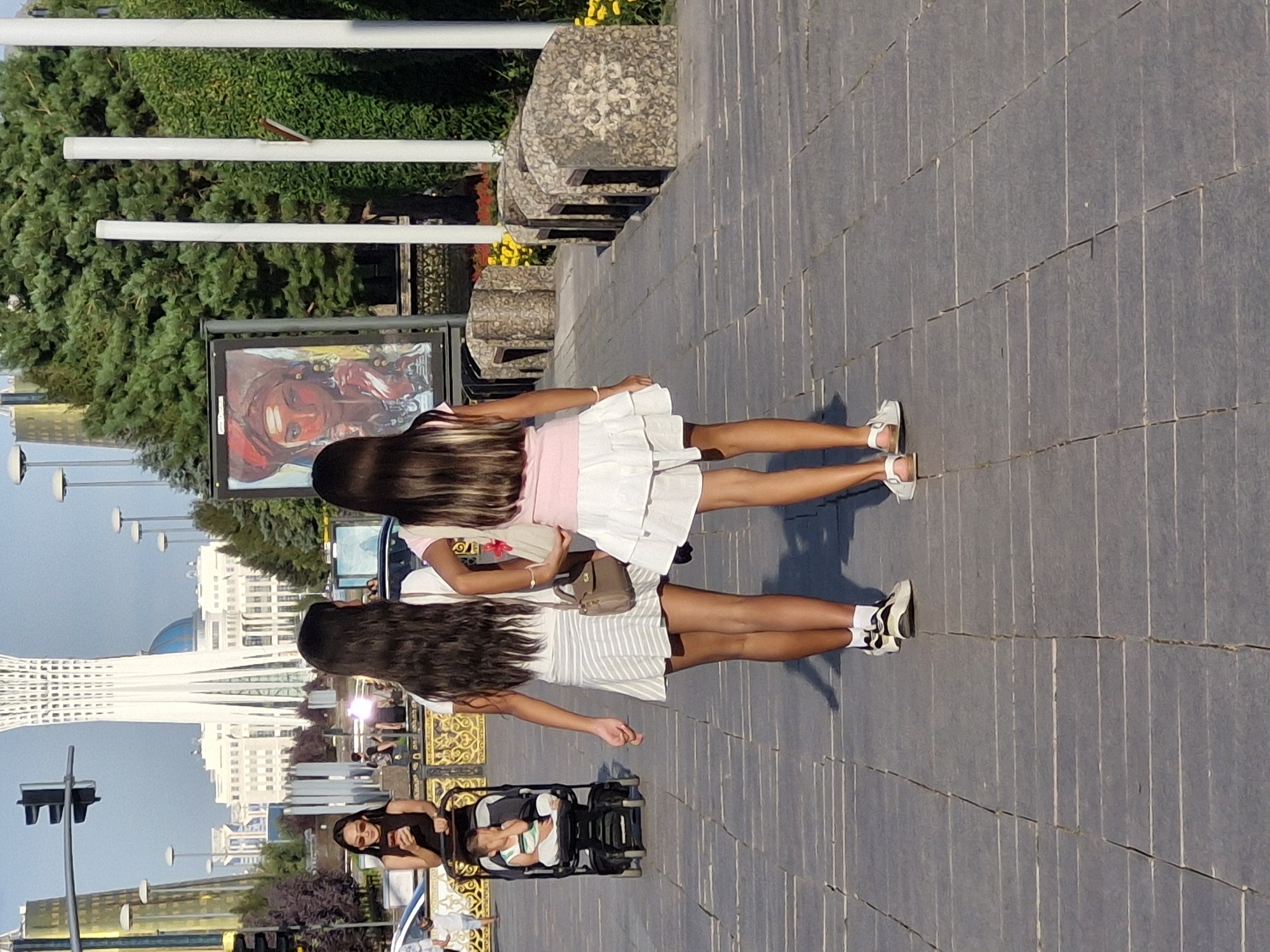 In the city of Astana, Kazakhstan, two young girls, viewed from behind, walk on a paved walkway. The girl on the left wears a white and grey striped skirt and a dark top. The girl on the right wears a white tiered skirt and a pink top. Both girls have long, dark hair. In the background, to the left, a woman in a dark top and sunglasses pushes a stroller containing a baby. Behind the woman, a large, white, futuristic building with a dome is visible, and to the right, a large poster of a woman's face in traditional attire is displayed. The environment appears to be an outdoor public space with trees and streetlights. The lighting suggests it is daytime.