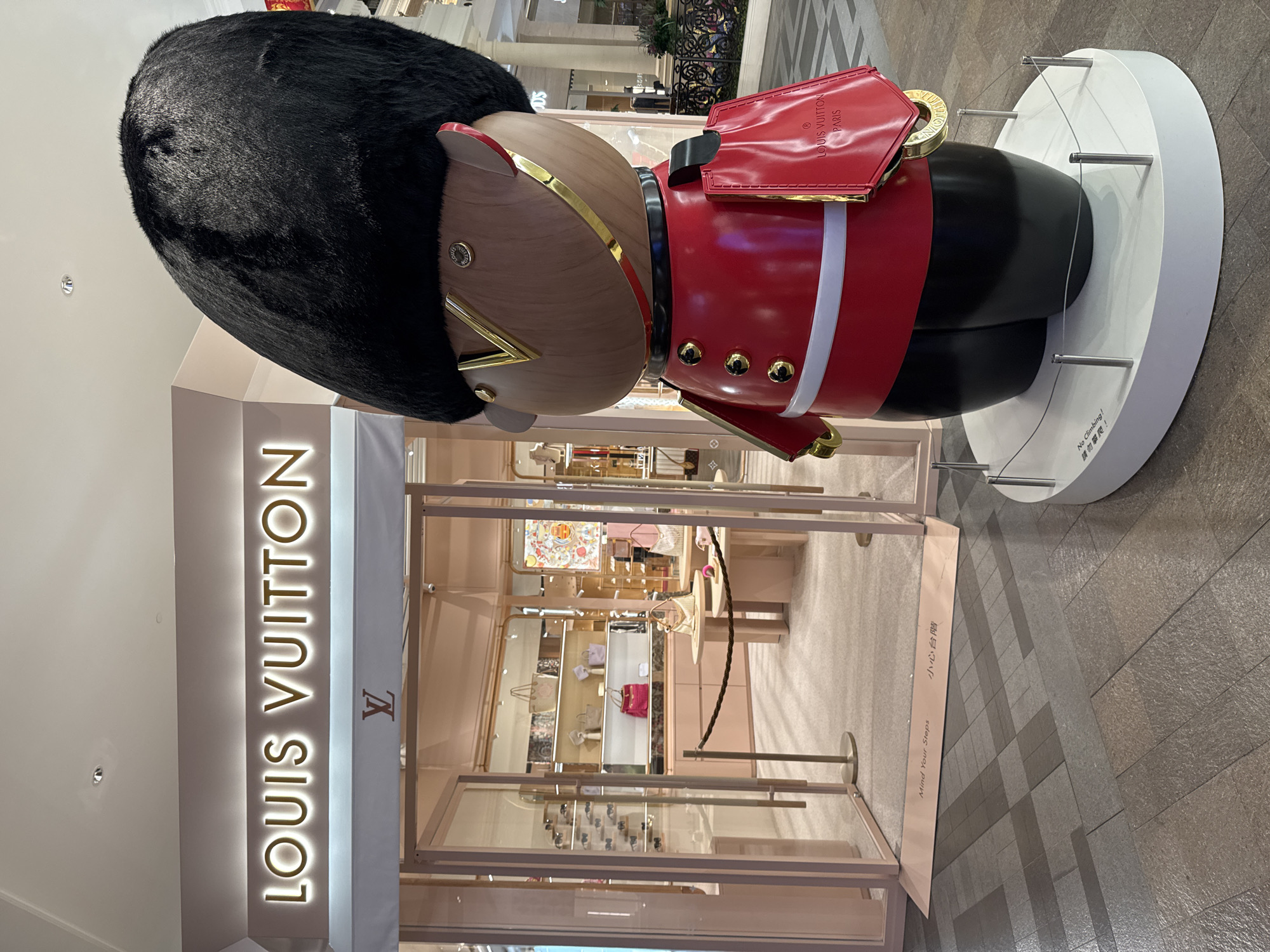 The image shows the front of a Louis Vuitton store in Unknown, Macao. The store's entrance is flanked by a large, stylized figurine resembling a British Guardsman, complete with a tall black fur hat and a red tunic with gold buttons. The figurine stands on a white circular base, with a warning sign in both English and Chinese indicating "No Climbing!" and "Mind Your Steps" at the store's entrance. The store's illuminated sign, "LOUIS VUITTON," is prominently displayed above the doorway. Through the glass entrance, shelves displaying various handbags and accessories are visible inside the store. The floor of the shopping mall is tiled with gray and brown stones.