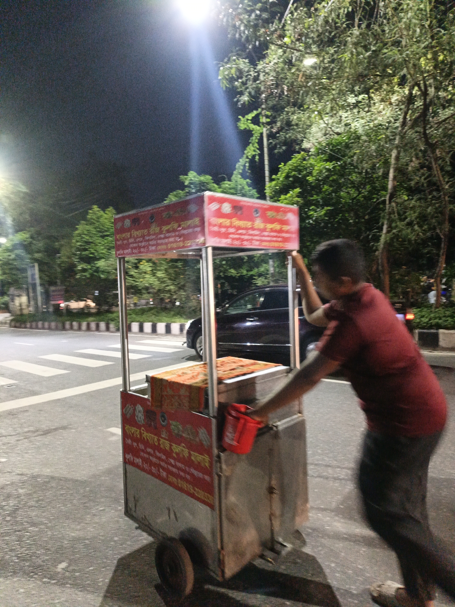 In this medium shot, a person wearing a maroon shirt and dark pants pushes a food cart with a red canopy down a street at night. The food cart has signage in a foreign language. The street has pedestrian crossings. In the background, trees and streetlights are visible, as well as a few cars. The scene appears to be in an urban environment.