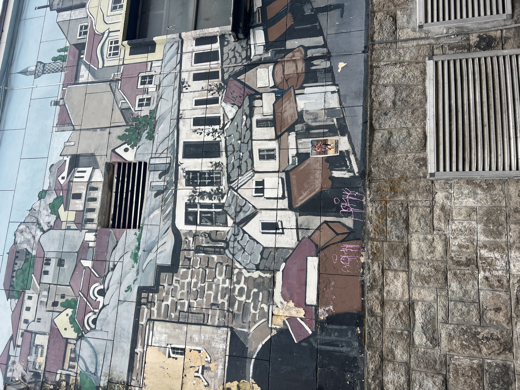 The image shows a wall mural in the city of Plovdiv, Bulgaria. The mural is painted on ceramic tiles and depicts a cityscape. The style of the mural is somewhat abstract, with simplified shapes and a limited color palette. The mural is partially obscured by two metal ventilation grates attached to the wall, as well as some graffiti. Adjacent to the mural is a section of wall constructed from large, irregular stone blocks.