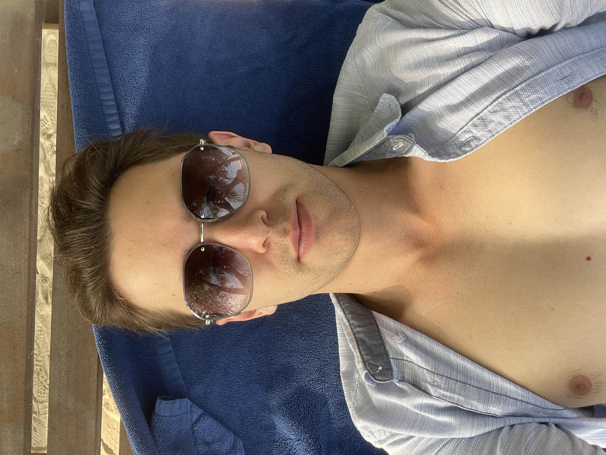A man is depicted lying down outdoors, seen from an overhead perspective. He is wearing silver-rimmed, dark sunglasses that reflect palm trees and bright sky, indicating a sunny, clear day, likely in a tropical or resort-like location. His dark hair is visible around his forehead, and he has light stubble on his face. His expression appears neutral and relaxed.
He is clad in a light blue, long-sleeved, collared shirt with a textured pattern, which is unbuttoned to reveal his bare chest. He is lying on a dark blue, terry cloth towel, which covers a surface that appears to be a sun lounger or similar outdoor seating. To the left side of the frame, next to his head, a wooden surface with a distinct grain is visible, possibly part of a deck or a wooden structure. The overall bright, natural lighting suggests it is daytime. No text is visible in the image.