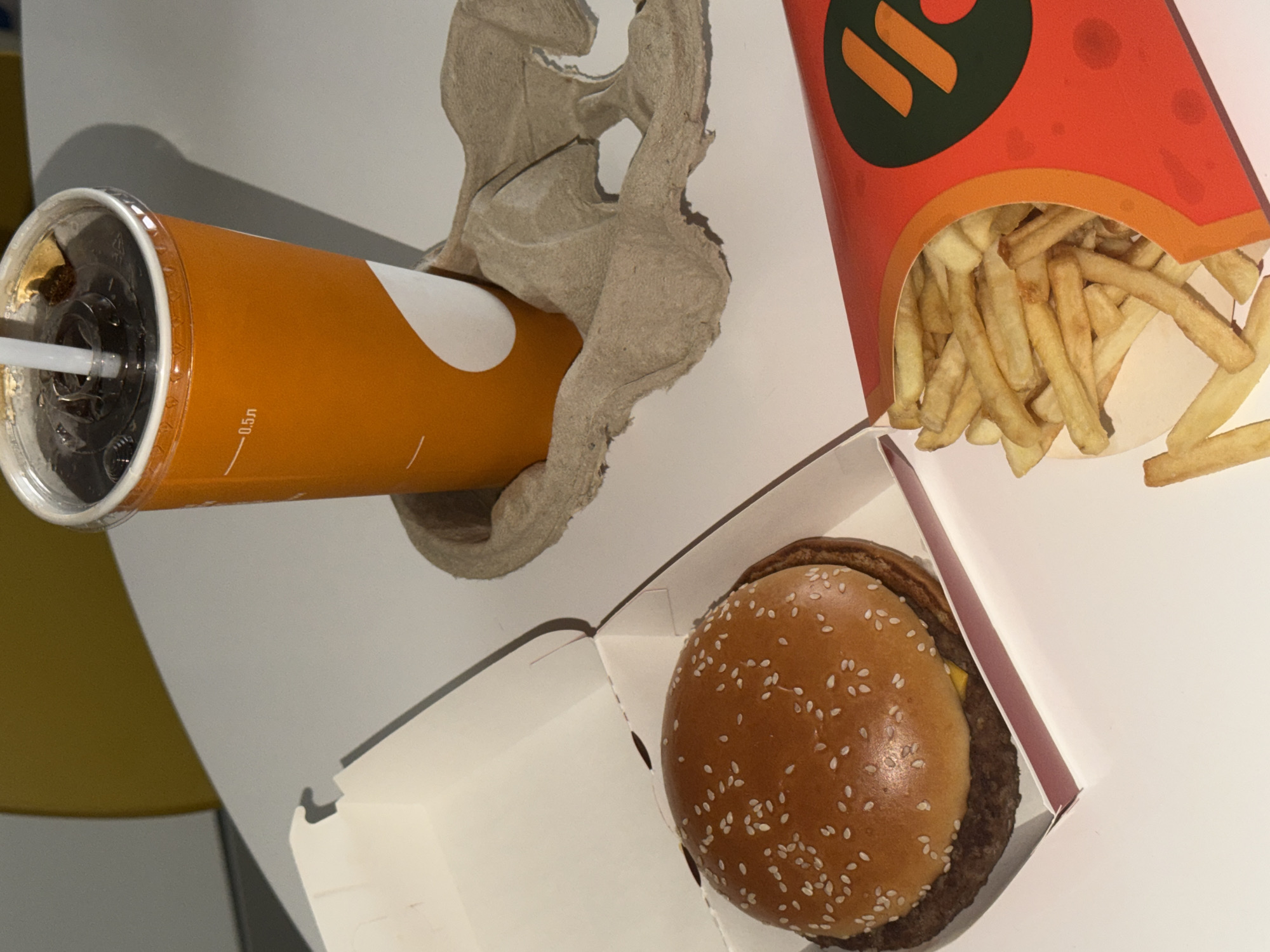 The media file displays a close-up, high-angle shot of a fast-food meal arranged on a clean, white table, likely indoors. The setup includes a beverage, a burger, and french fries, all presented in their respective brand packaging.
On the left side of the frame, a large, orange-colored soda cup with a white, stylized wave pattern is visible. The cup is fitted with a clear plastic lid and a white straw, revealing a dark beverage inside. Text on the cup clearly indicates "0.5л," signifying a 0.5-liter volume. This cup is supported by a light brown, molded cardboard carrier, typically used for transporting drinks.
To the right, a classic burger is placed in an open, white cardboard box. The burger features a sesame seed bun, a visible beef patty, and a slice of melted cheese. Next to the burger, a red and orange carton overflows with golden-brown french fries. The fries carton is adorned with a prominent logo consisting of abstract orange shapes set against a dark green background on a red field. Given the location context of Khimki, Russia, this logo is identifiable as belonging to "Vkusno i Tochka," the Russian fast-food chain successor to McDonald's.
The overall scene suggests a recently served meal, possibly for a customer in a restaurant or as a take-out order. The lighting is bright and even, indicating an indoor environment. A blurred yellow surface is marginally visible in the extreme background on the left, hinting at the surrounding decor. No people or discernible emotions are visible.
