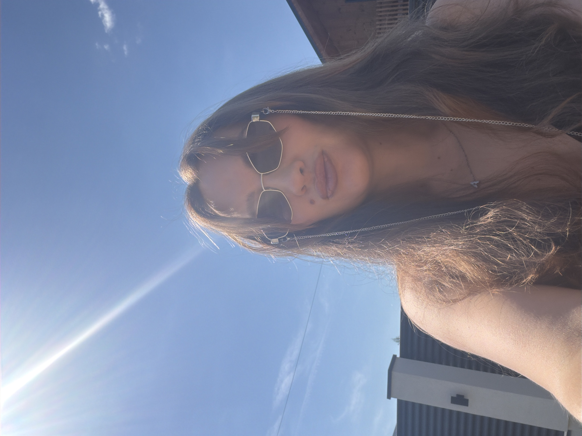 A woman with long brown hair is taking a selfie outdoors on a sunny day. She is wearing sunglasses with a chain and a necklace with a small pendant. In the background, there is a blue sky with a bright sunbeam, a fence, and a wooden building. The location is identified as Malinovka, Russia.