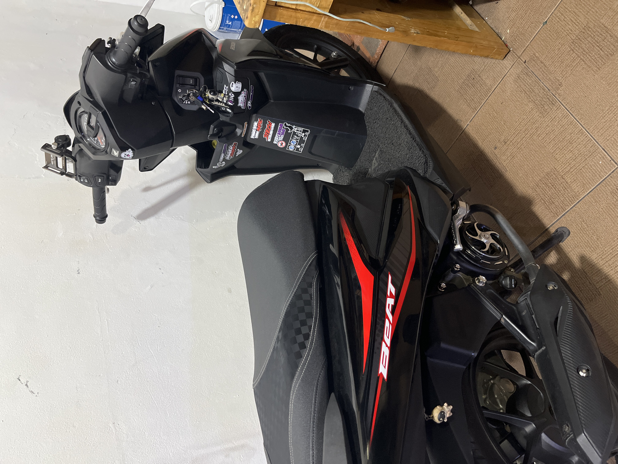 A black Honda Beat scooter is parked indoors, likely in a residential area or garage, near a white wall and a wooden structure. The scooter has several stickers on its body, including one with the word "BEAT" in red and white. A small, tan-colored keychain figure is attached to the left side of the scooter. The flooring appears to be tiled. The scooter is upright, supported by its kickstand, and is not in motion. There are no people visible in the image. The lighting is artificial, suggesting an indoor environment. The overall scene is static and focuses on the parked scooter.