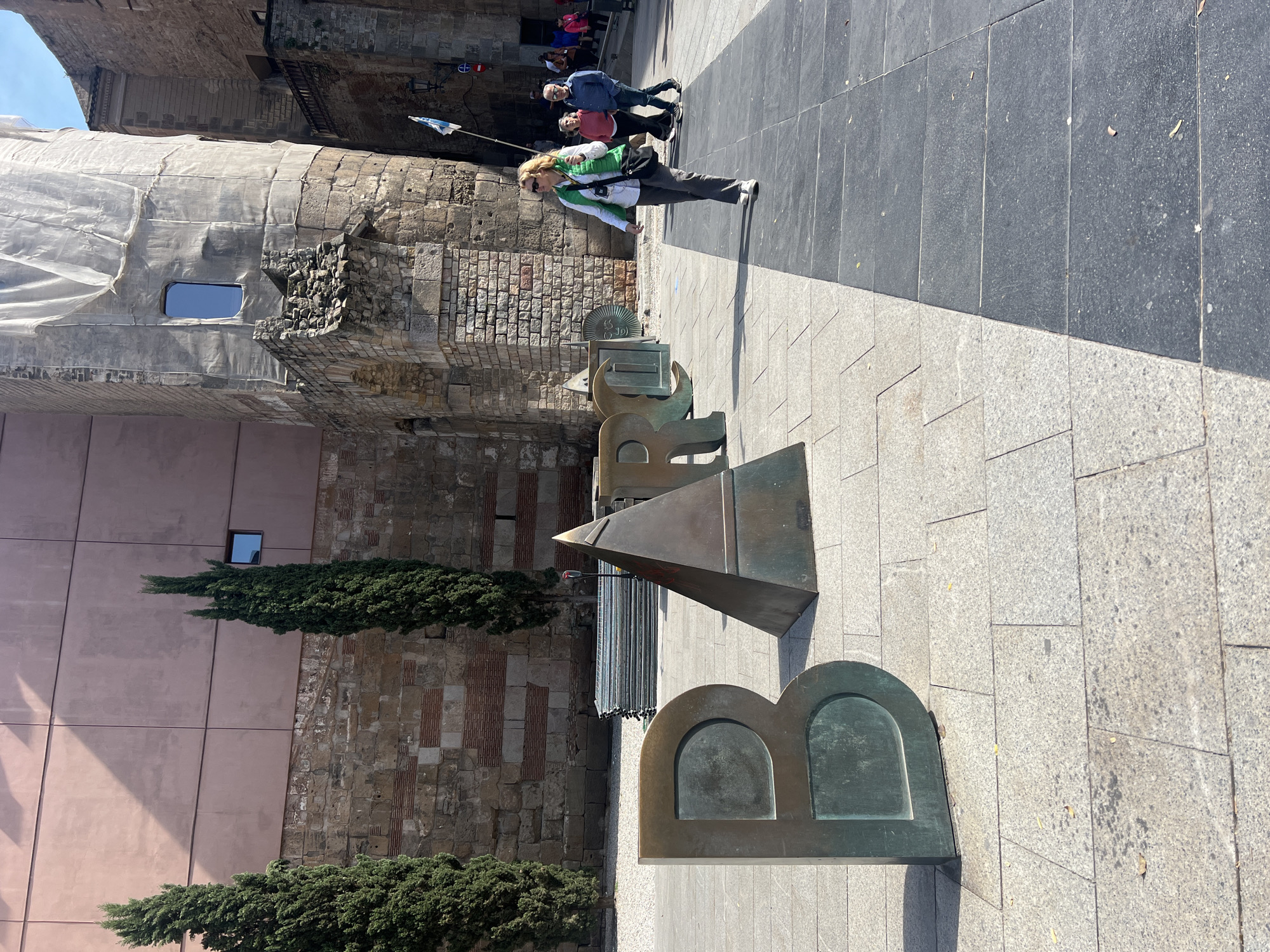 The image captures a street scene in Barcelona, Spain, featuring a large bronze sculpture spelling out "BARC" with the partial letters of "BARCELONA" on the ground. A woman is walking to the right, along a cobblestone street, with several other people farther in the background. The sculpture is beside an old stone and brick building. The lower building is pink with two trees against it. The top part of the building has netting covering part of it. The pavement changes to dark tile near the top of the frame.