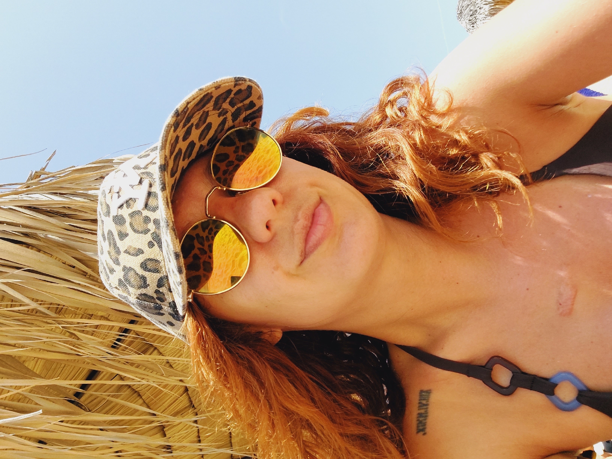 A woman with long, wavy red hair is pictured from the chest up. She is wearing a leopard-print baseball cap and round, gold-rimmed sunglasses with reflective orange lenses. Her head is tilted slightly to her left, and she is looking towards the camera with a slight smile. She is wearing a black bikini top with blue and black rings on the strap. A Roman numeral tattoo, "XXIV.IV.", is visible on her left shoulder. A small, light-colored scar is present on her upper chest. The background consists of a thatched umbrella providing shade and a clear blue sky. The image appears to be taken outdoors, possibly at a beach or resort.