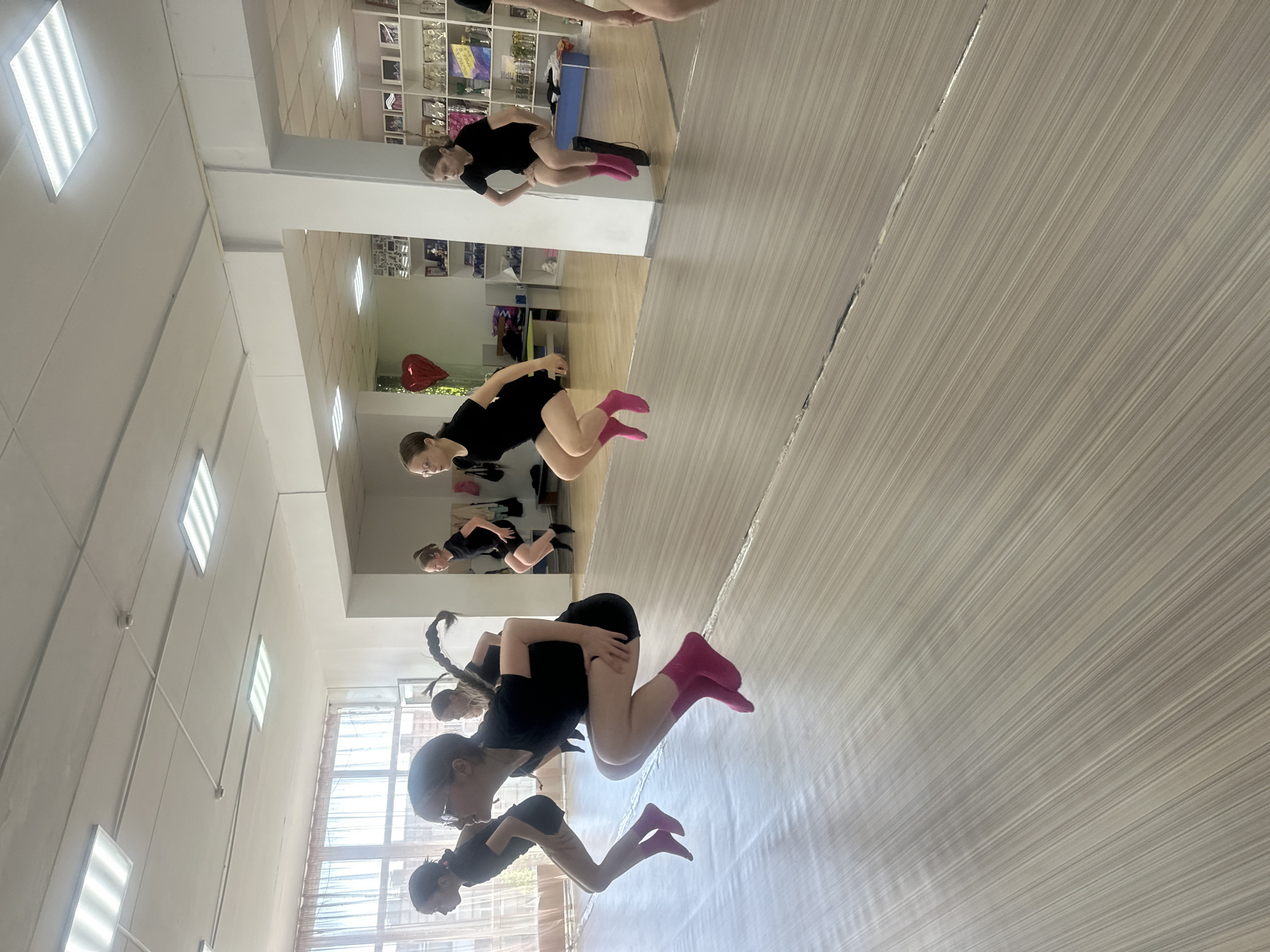 The image shows a group of young women in a dance studio in Votkinsk, Russia. The studio has light-colored walls, and a wood-look floor. One wall is mirrored, reflecting the scene. The dancers are wearing black dance attire and bright pink socks, posed in synchronized motion. The fluorescent lights are visible in the ceiling. Shelves with various items are visible in the reflection of the mirror. A red heart shaped balloon is also visible in the reflection.