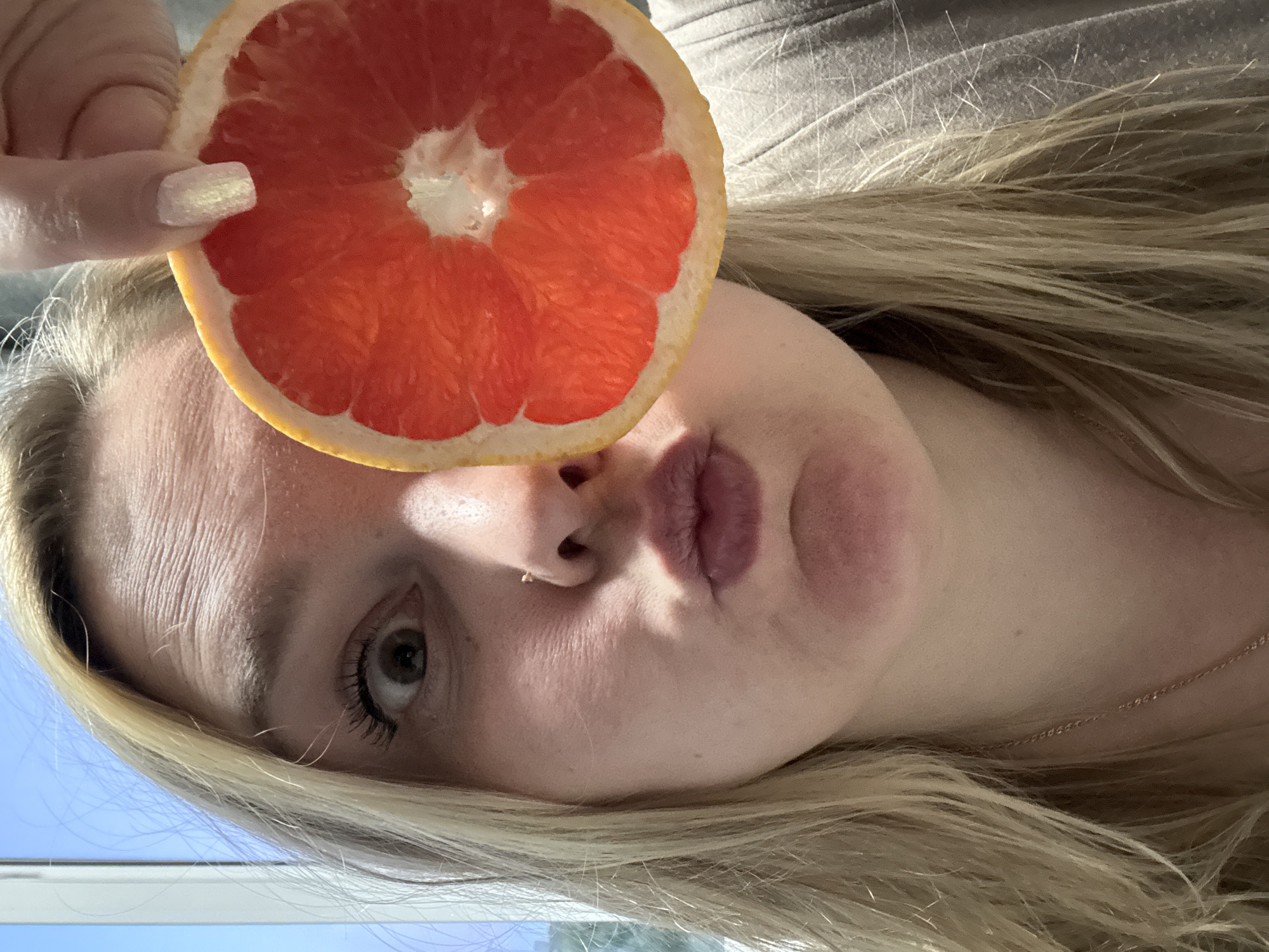 A young, fair-skinned woman with blonde hair is holding a slice of grapefruit up to her forehead, with her index finger partially visible near the grapefruit. She is looking slightly downward. A delicate chain necklace is visible around her neck. The image appears to have been taken indoors, with a light blue section of what might be a window or wall visible in the lower left corner. Without further context, determining if this image originated in the specific location of Марковский, Russia is not possible based on only the image itself. There are no landmarks or features within the image to corroborate that location.