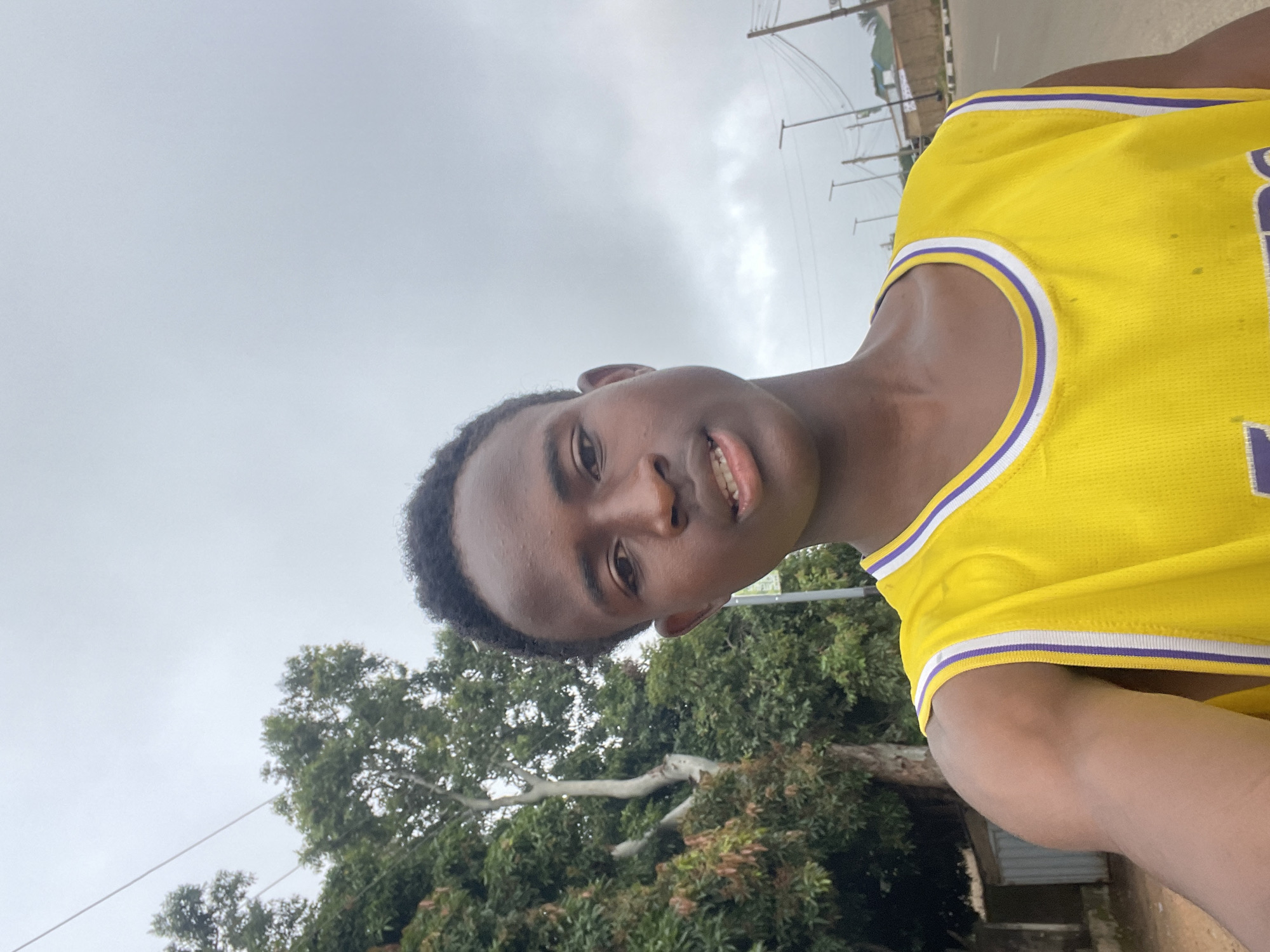 The image is a selfie taken outdoors, likely on a cloudy day, possibly in the city of Jos, Nigeria. In the foreground, a young Black male is centered, wearing a bright yellow basketball jersey with white and purple trim around the neckline and armholes. His skin is dark, and his hair is short and dark. He is looking directly at the camera with his mouth slightly open, revealing his teeth. To the left of the subject, there is lush green foliage from trees, with a large tree trunk visible. Behind the trees and to the right of the subject, a paved road is visible, with streetlights and utility poles lining the side. Some buildings are also faintly visible in the background on the right side of the road. The overall environment suggests a semi-urban or suburban setting.